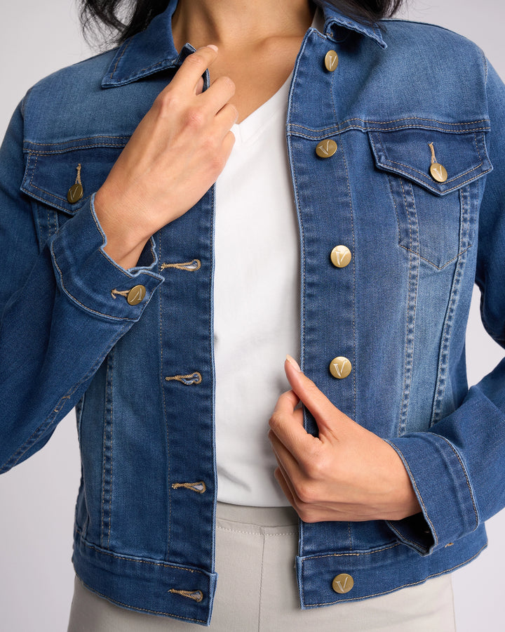 A person is buttoning the Vassalli Ratio Jacket - Blue Wash Denim over a white shirt, with one hand by the collar and the other by the bottom buttons. Their face is not visible.