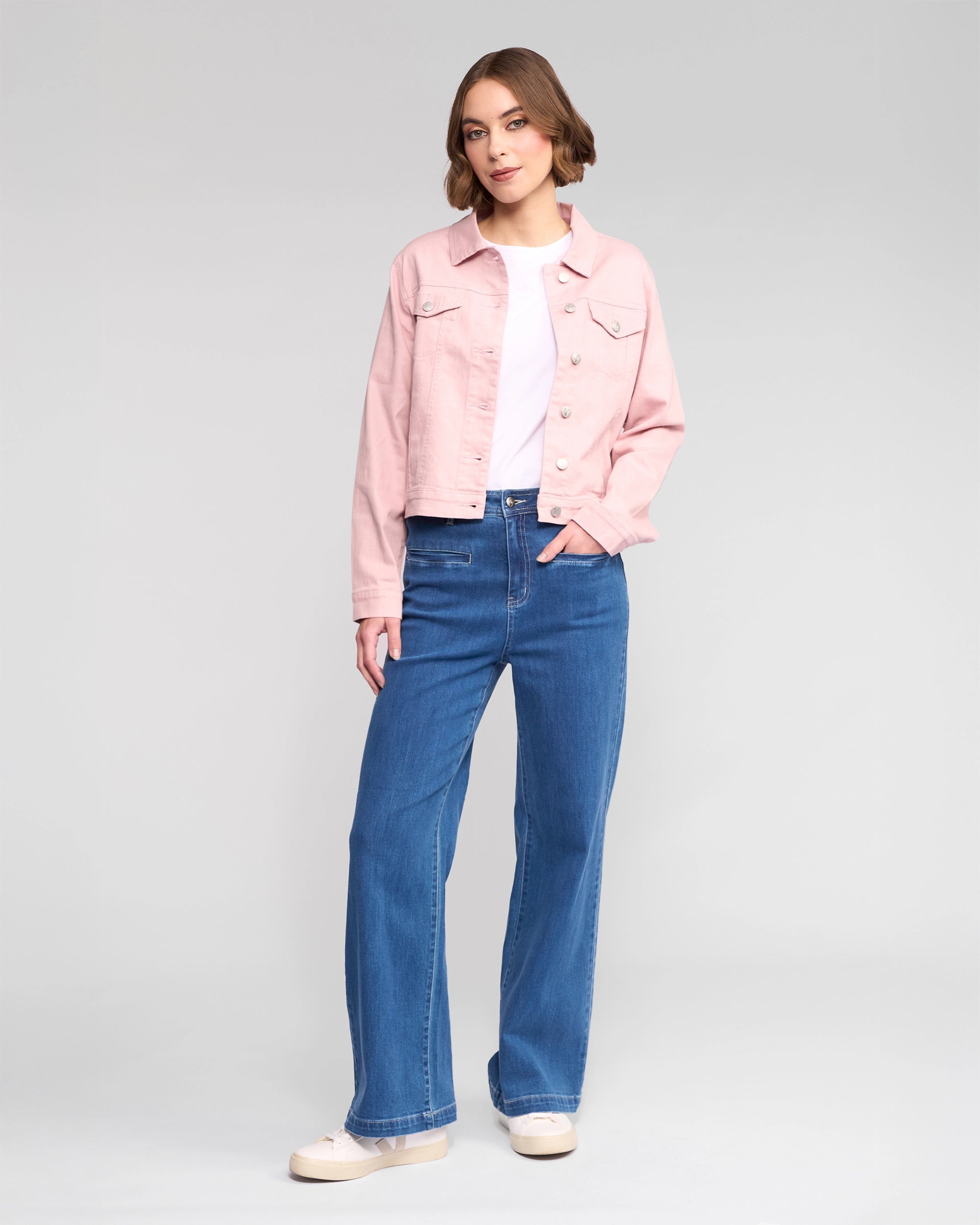 A woman with short brown hair stands against a plain background wearing the Vassalli Ratio Jacket in French Rose Denim over a white top, blue wide-leg jeans, and beige shoes, one hand in her pocket for an effortlessly chic look.