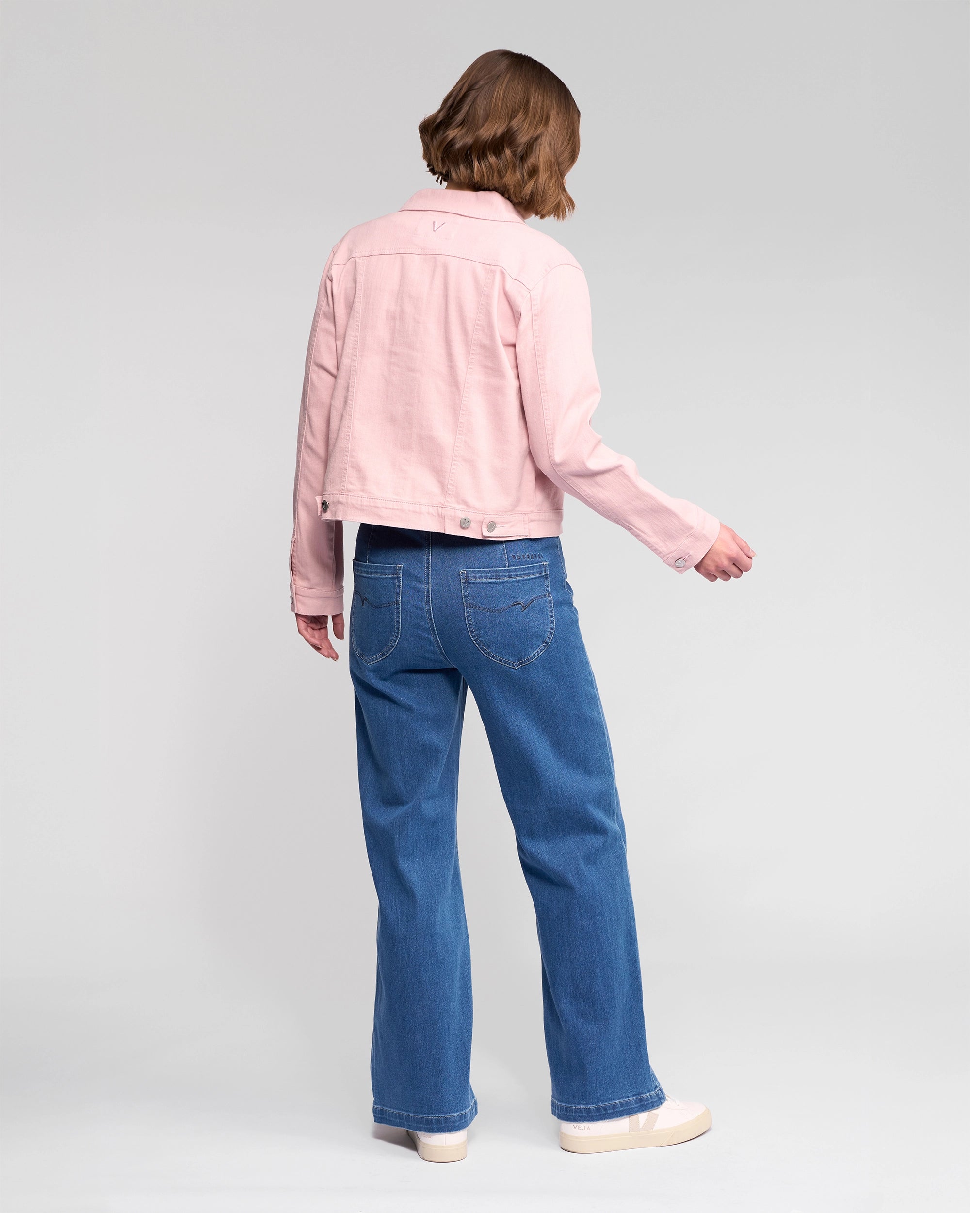 A person with short, light brown hair stands with their back to the camera, wearing the Vassalli Ratio Jacket in French Rose Denim, blue stretch denim jeans, and light-colored platform shoes against a plain white background.