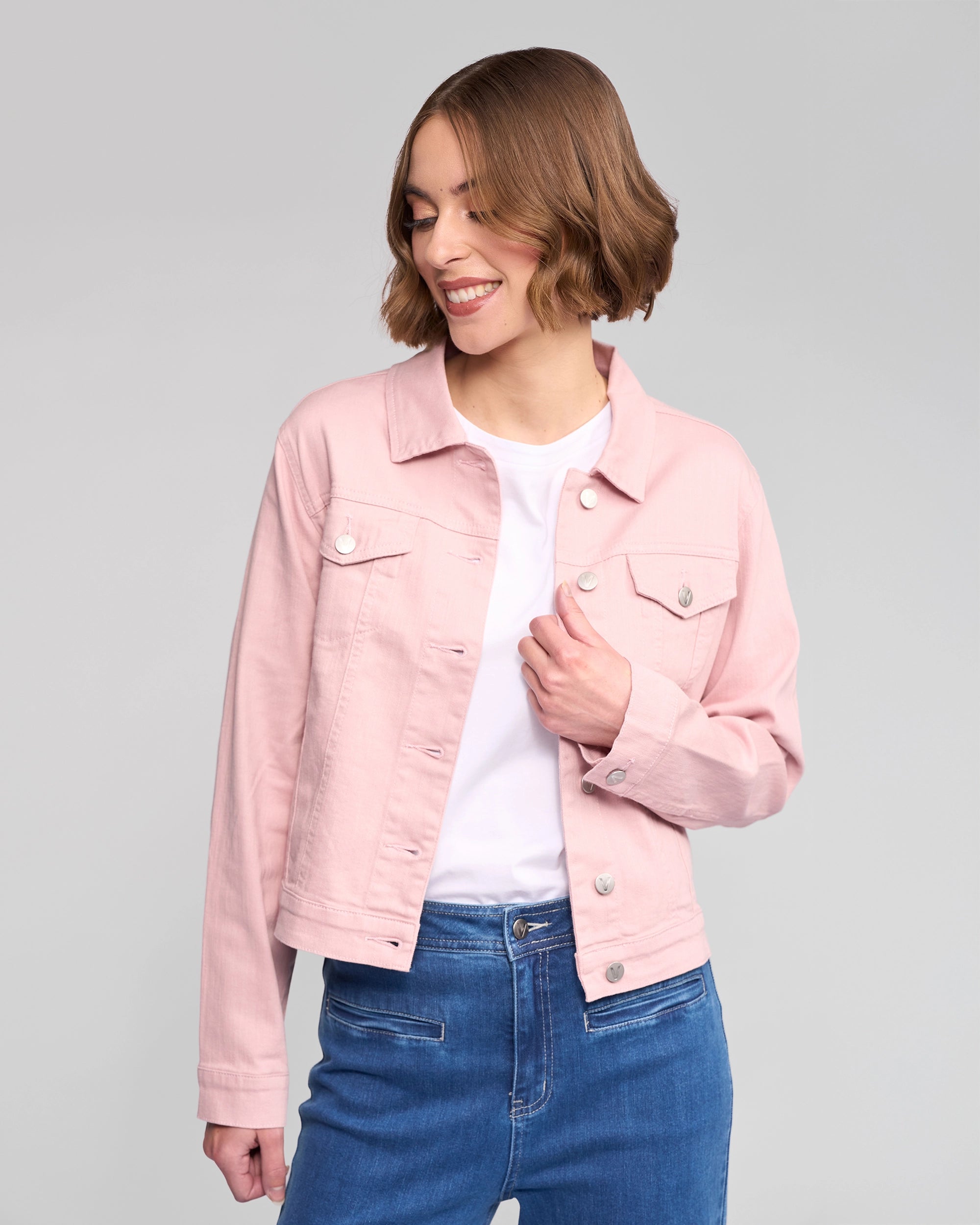 A woman with short brown hair smiles while wearing the Vassalli Ratio Jacket in French Rose Denim over a white shirt and blue jeans against a plain light gray background.
