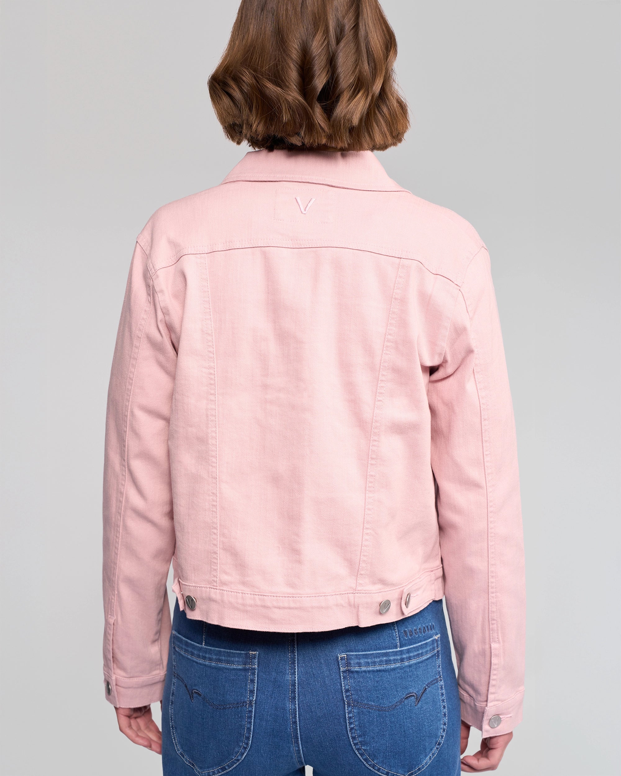 A person with short, wavy brown hair is seen from behind wearing the Vassalli Ratio Jacket in French Rose Denim and blue jeans against a plain gray background.