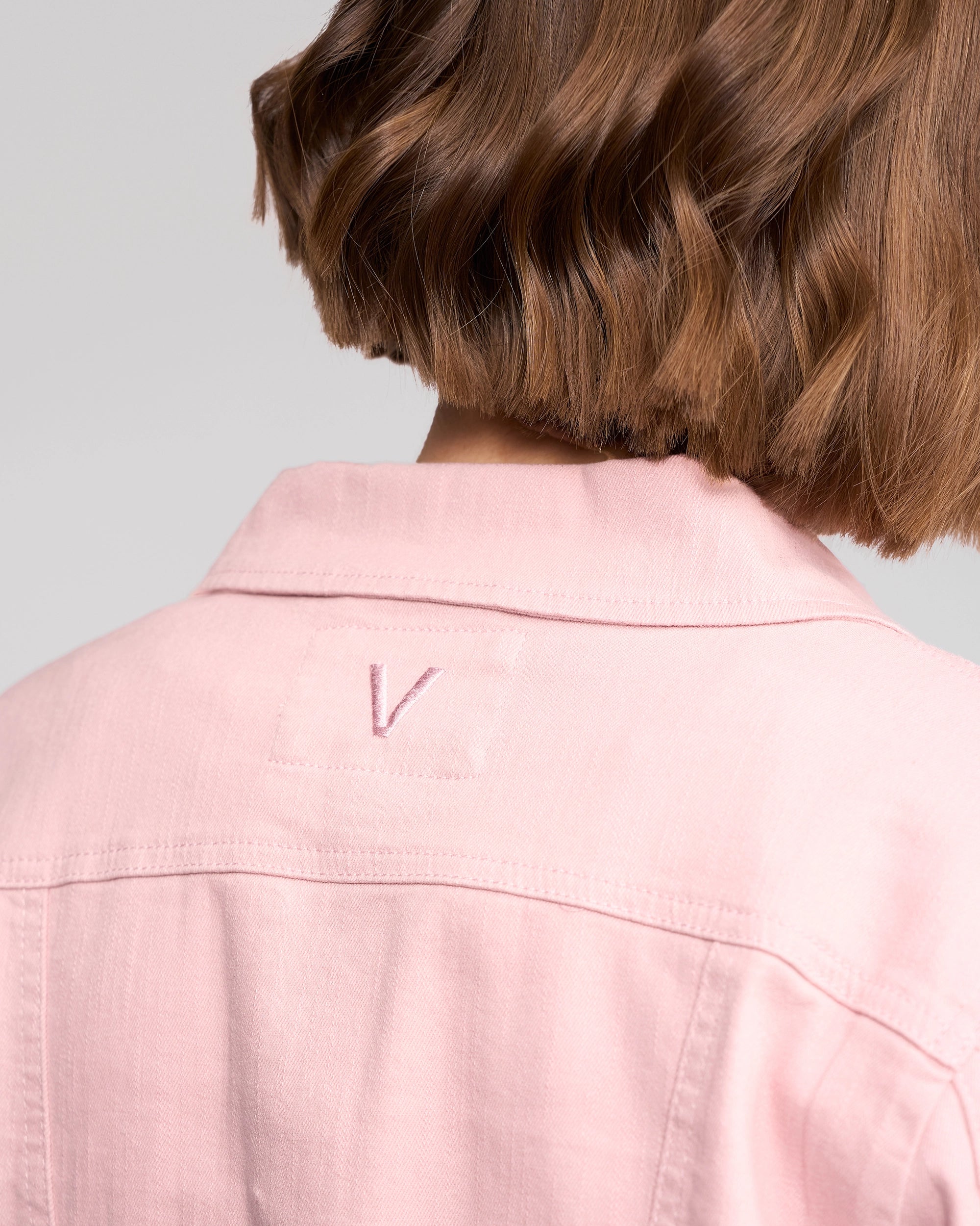 A person with short, wavy brown hair is shown from behind, wearing the Vassalli Ratio Jacket - French Rose Denim, which features a relaxed fit, subtle stretch denim, and a stitched V detail on the upper back.