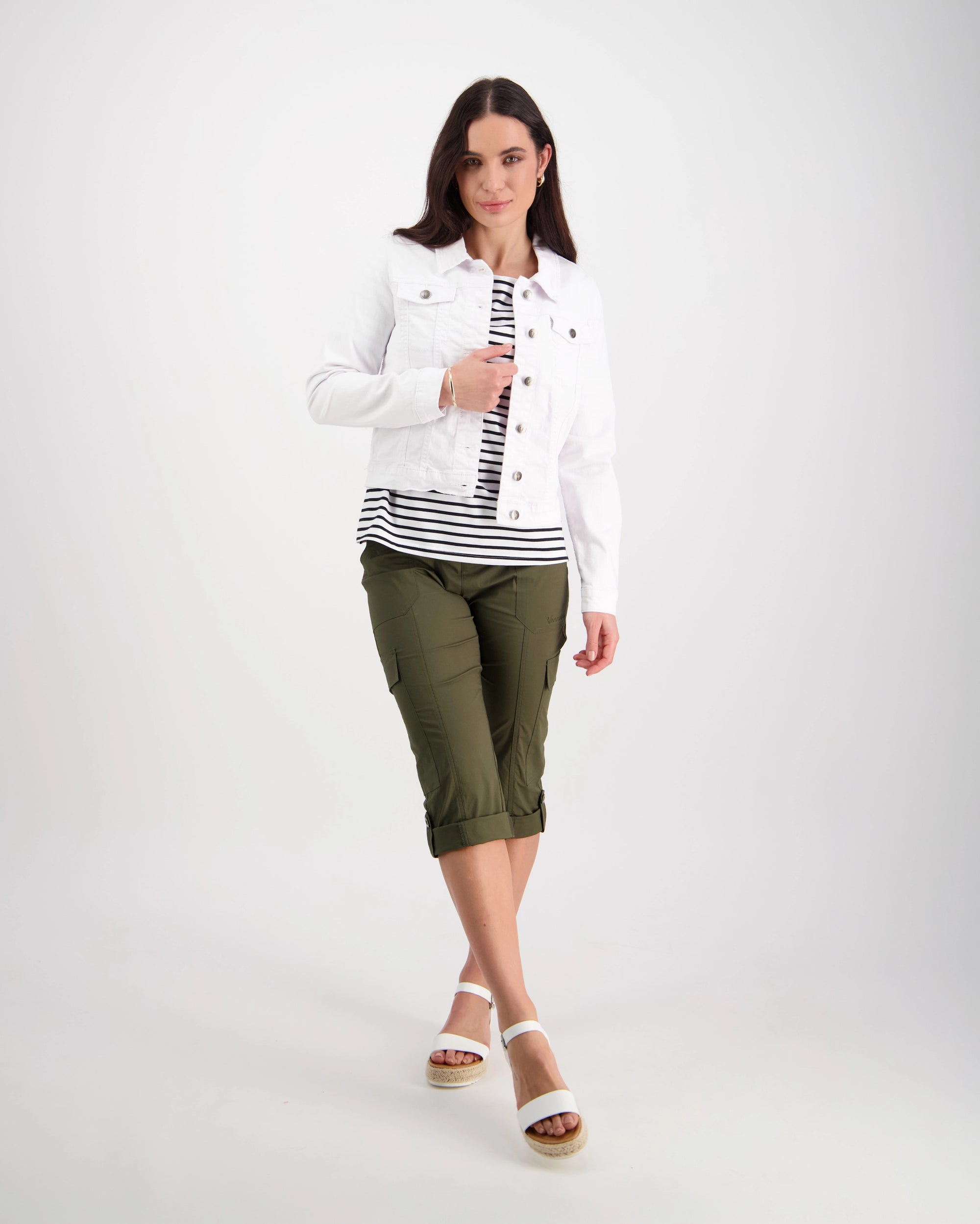 A woman with straight brown hair and a relaxed expression stands against a white background, wearing the Vassalli Ratio Jacket in white denim over a striped top, olive green cropped pants, and white sandals, with one hand in her pocket.