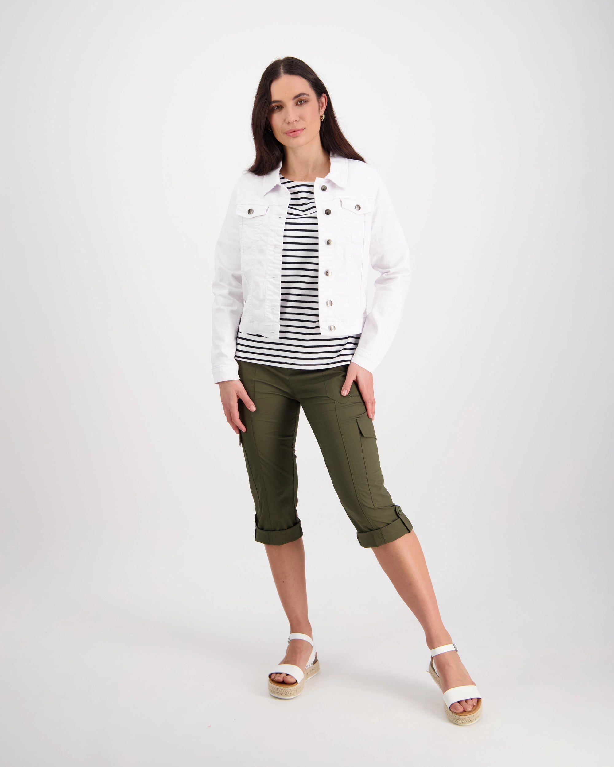 A woman with straight dark hair wears the Vassalli Ratio Jacket in white denim over a black and white striped shirt, paired with olive green capri pants and white platform sandals, posing against a plain white background.