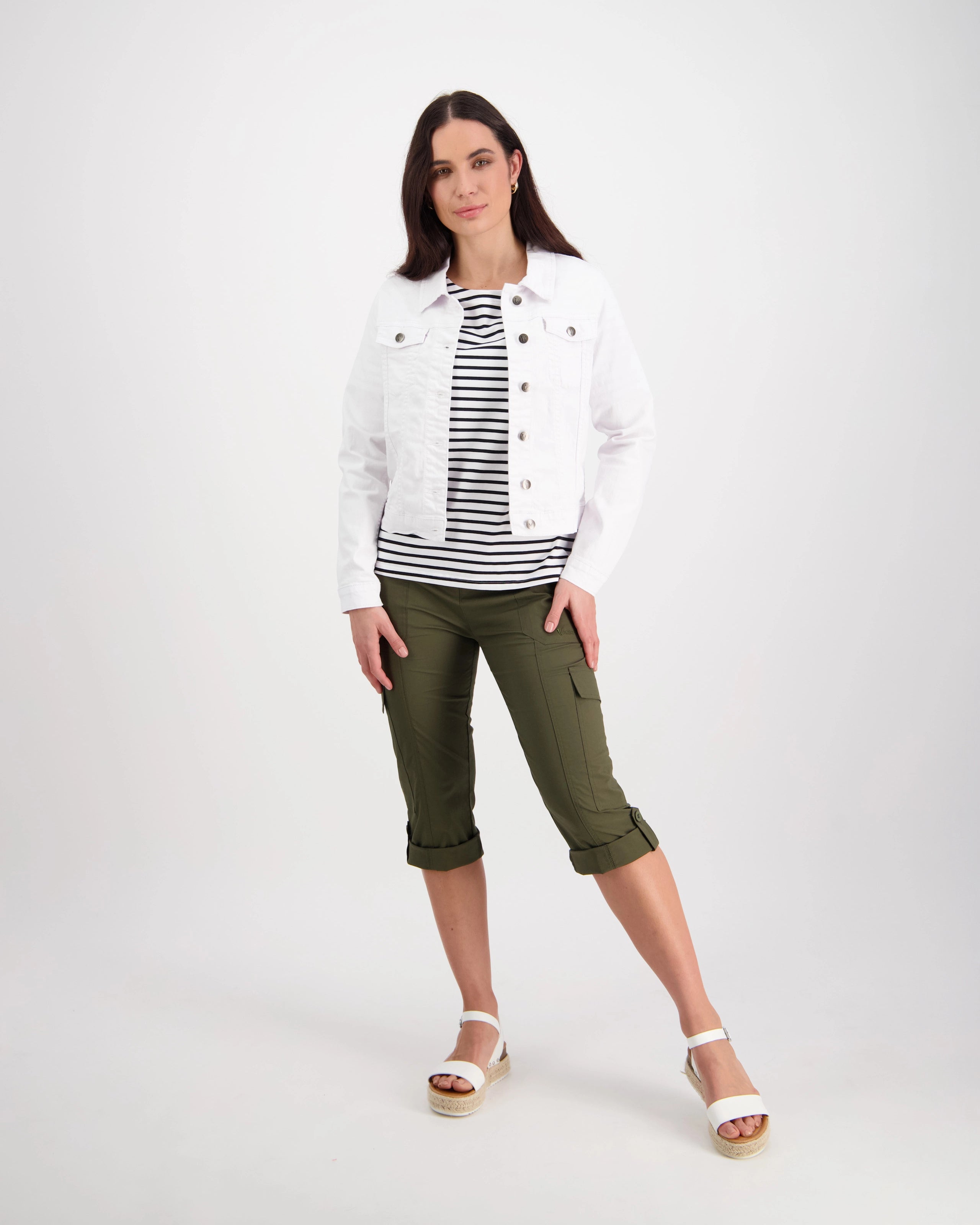A woman with straight dark hair wears the Vassalli Ratio Jacket in white denim over a black and white striped shirt, paired with olive green capri pants and white platform sandals, posing against a plain white background.