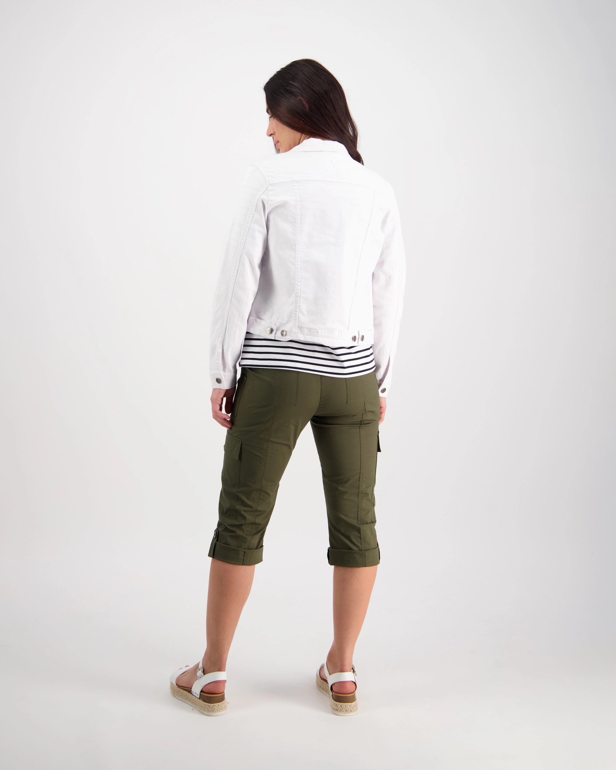 A woman stands with her back to the camera, wearing the Vassalli Ratio Jacket in white denim over a striped shirt, paired with relaxed fit olive green cropped cargo pants and white sandals, all set against a plain white background.