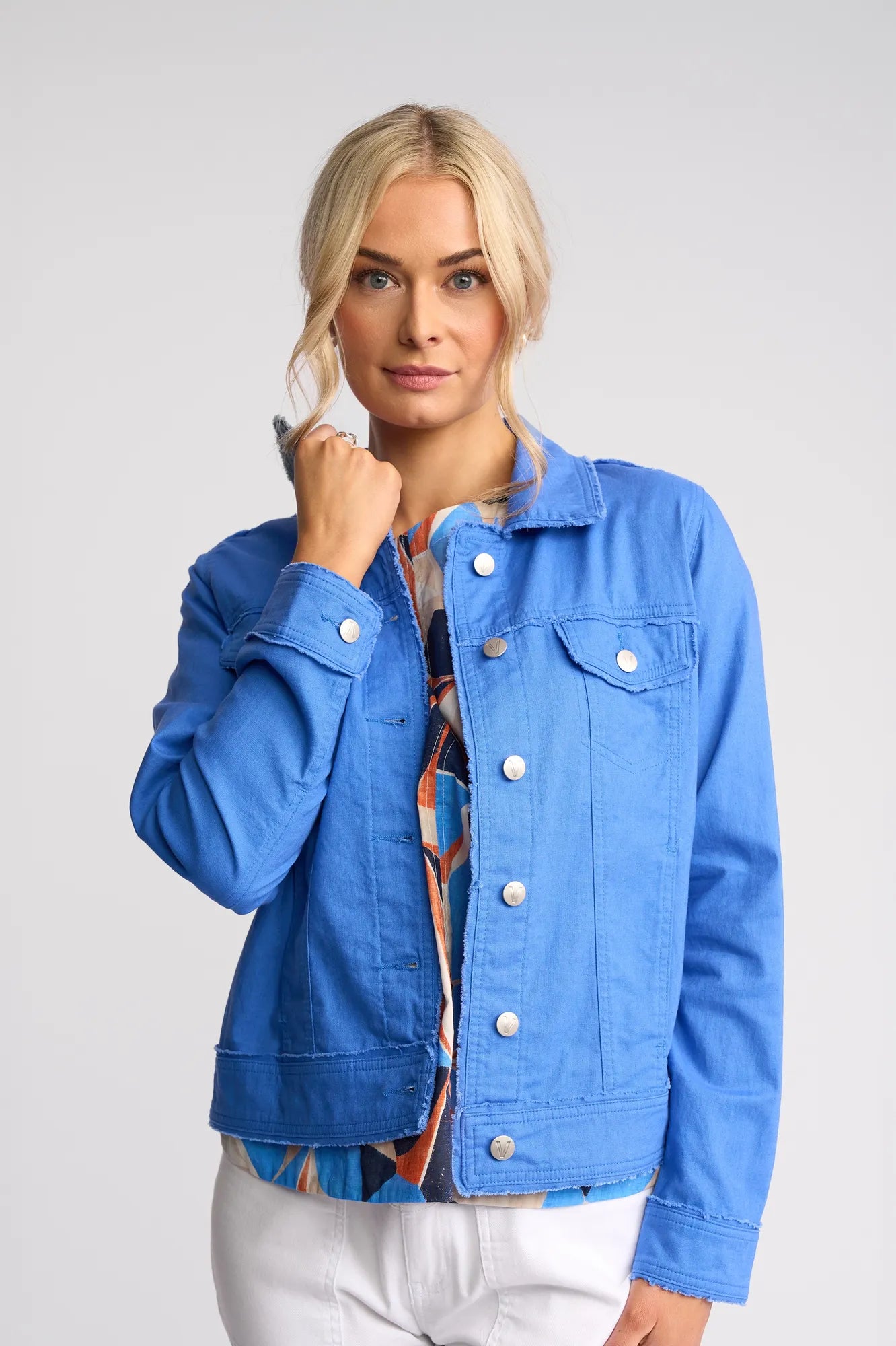 A woman with blonde hair wears the Vassalli Aura Jacket in French Blue, crafted from a soft linen-cotton blend, layered over a colorful patterned shirt and white pants, standing against a plain light gray background.