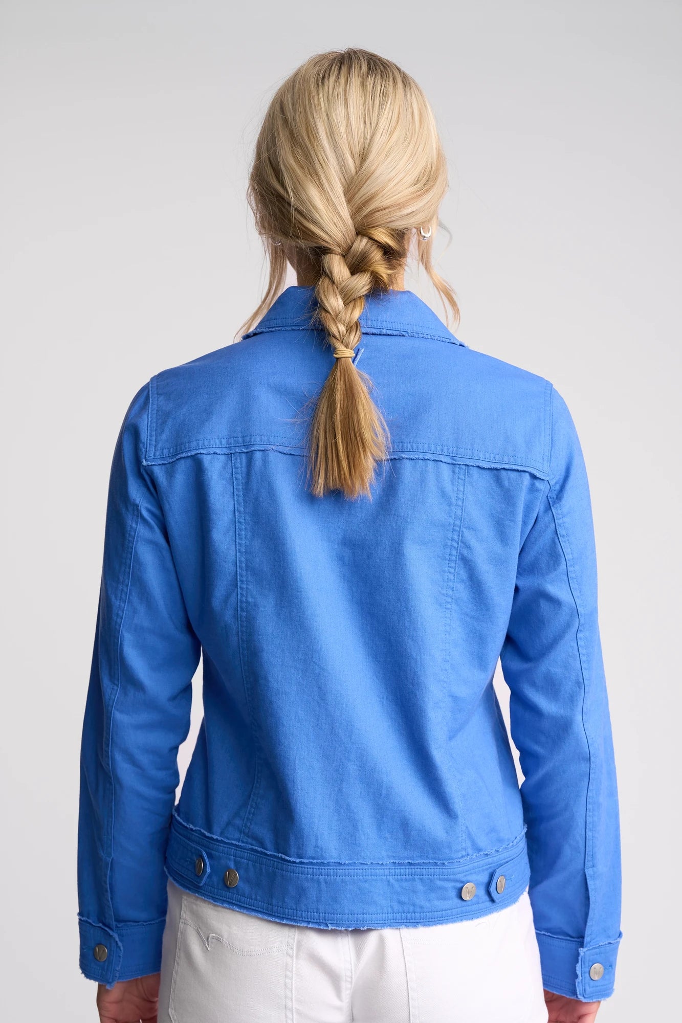 A woman with blonde hair in a braid is shown from behind, wearing the Vassalli Aura Jacket in French Blue—a soft linen cotton blend—paired with white pants, standing against a plain light background.