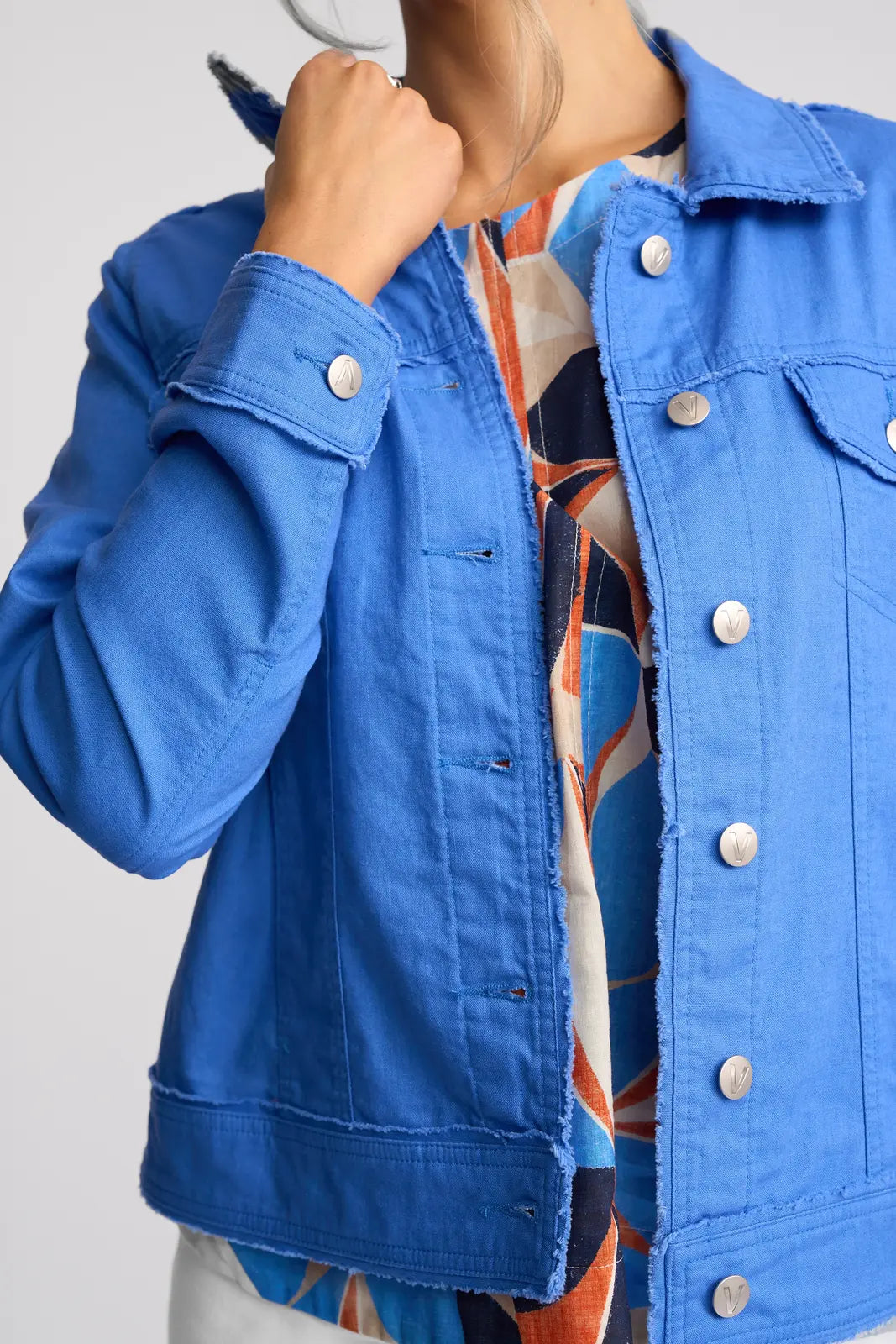 A person wearing the Vassalli Aura Jacket in French Blue, crafted from a linen-cotton blend, over a colorful abstract-patterned top, holding the jacket collar with one hand. The face is partially out of frame.
