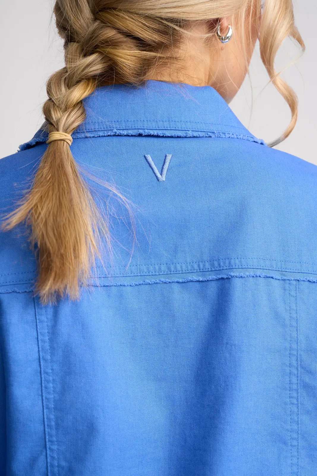 A person with blond hair in a braid is seen from behind wearing the Vassalli Aura Jacket in French Blue, made from a linen cotton blend and featuring a small, embroidered V below the collar.