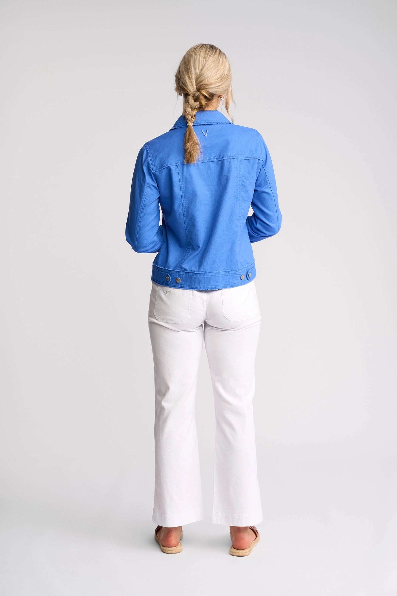 A woman with blonde hair in a braid, wearing the Vassalli Aura Jacket in French Blue—a linen cotton blend—paired with white pants, stands facing away in a studio with a plain white background.