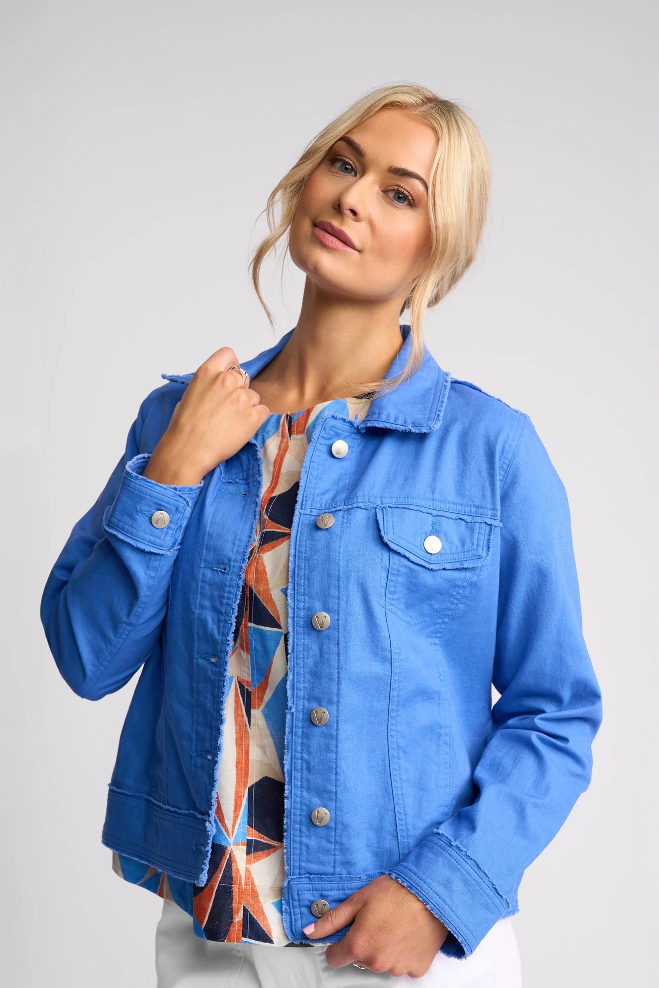 A woman with blonde hair wears the Vassalli Aura Jacket in French Blue, crafted from a linen-cotton blend, layered over a colorful patterned top, as she poses against a plain light background.