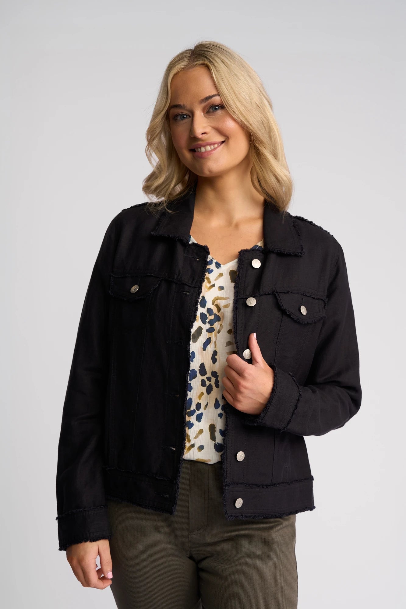 Smiling woman with blonde hair wearing a Vassalli Aura Jacket in navy—an elegant button-up style crafted from a viscose linen blend—over a patterned top and olive pants, standing against a plain light background.