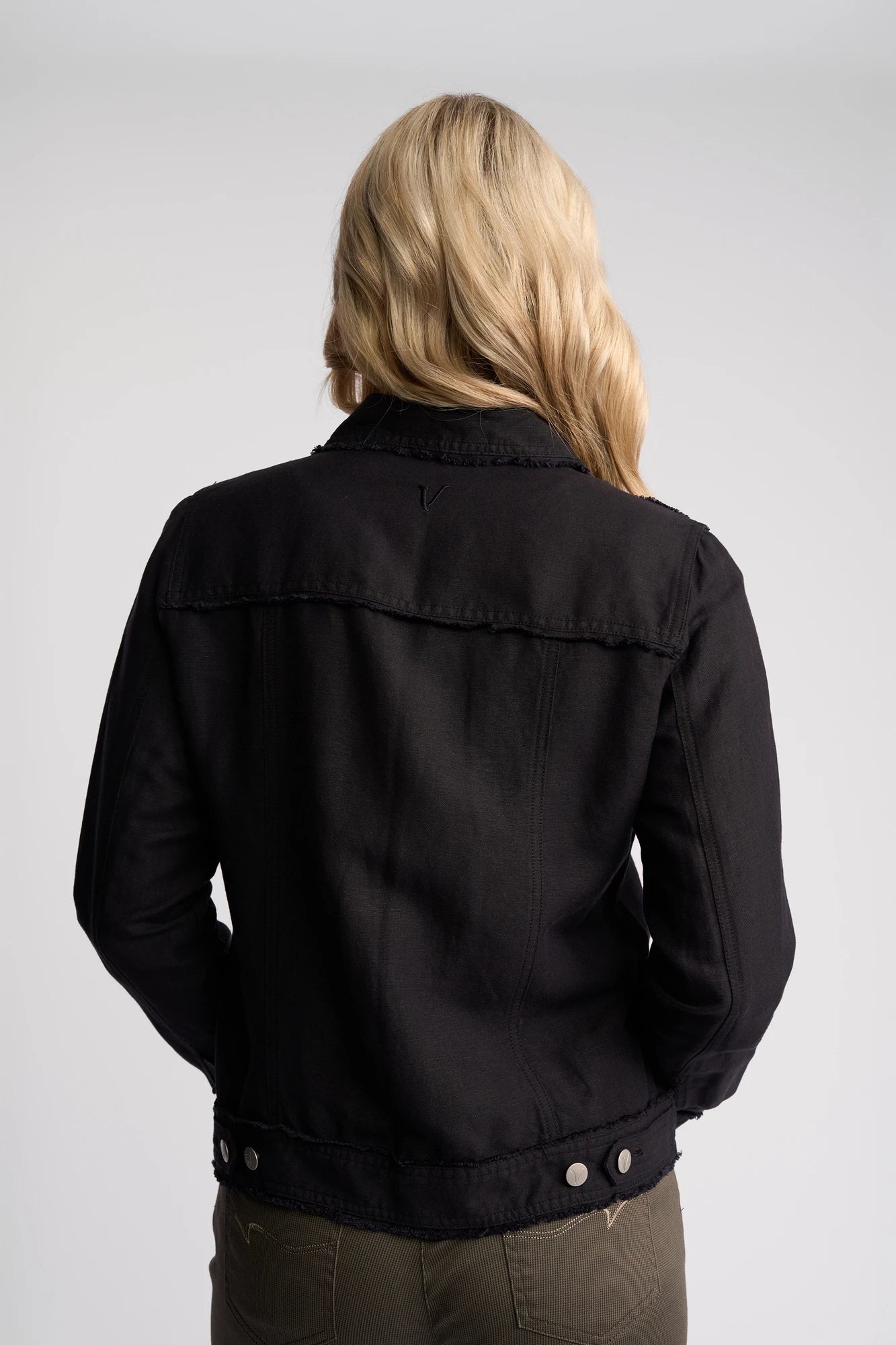 Seen from behind against a plain light gray background, a person with long blonde hair is wearing the Vassalli Aura Jacket in Navy paired with olive green pants.