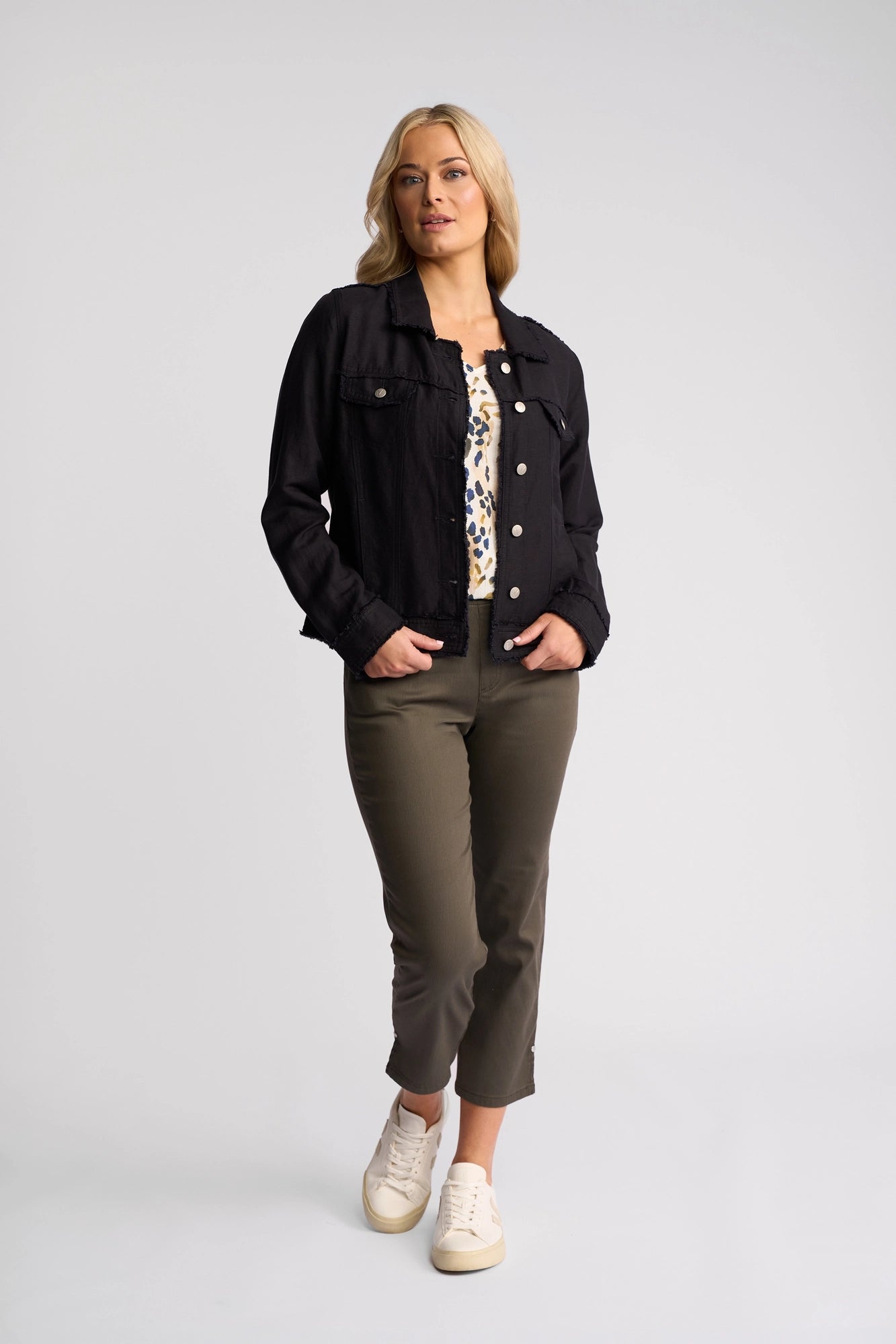A woman stands with hands in her pockets, wearing the Aura Jacket by Vassalli—a stylish navy denim jacket—paired with a printed top, olive cropped pants made from a soft viscose linen blend, and white sneakers, against a plain light background.
