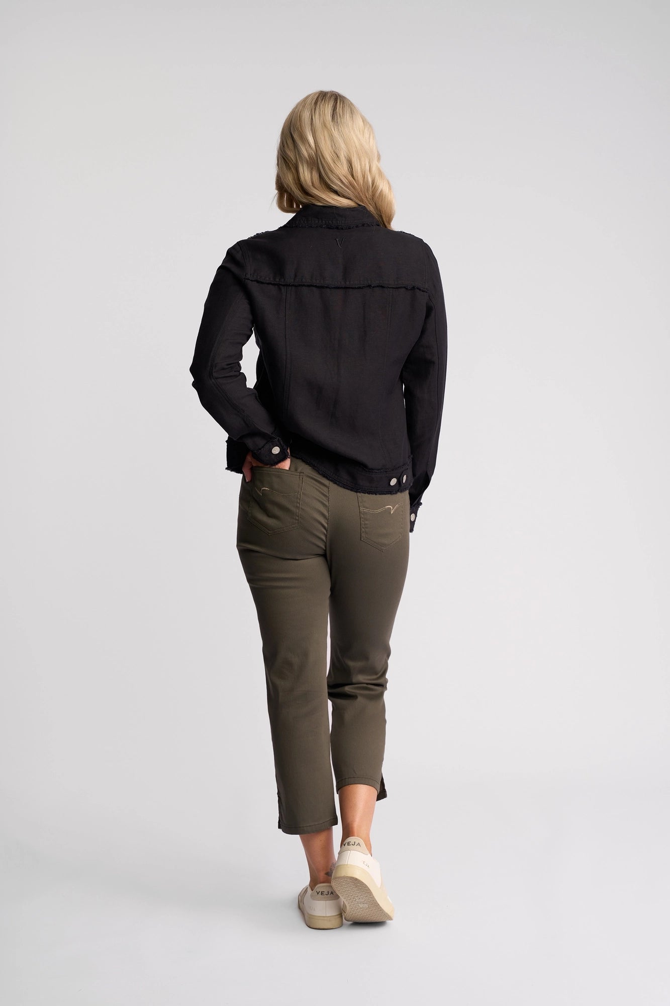 A person with blonde hair, wearing a black long-sleeve shirt, olive green pants, and white sneakers, stands facing away from the camera against a plain white background, showcasing the relaxed silhouette of the Vassalli Aura Jacket - Navy in a soft viscose linen blend.