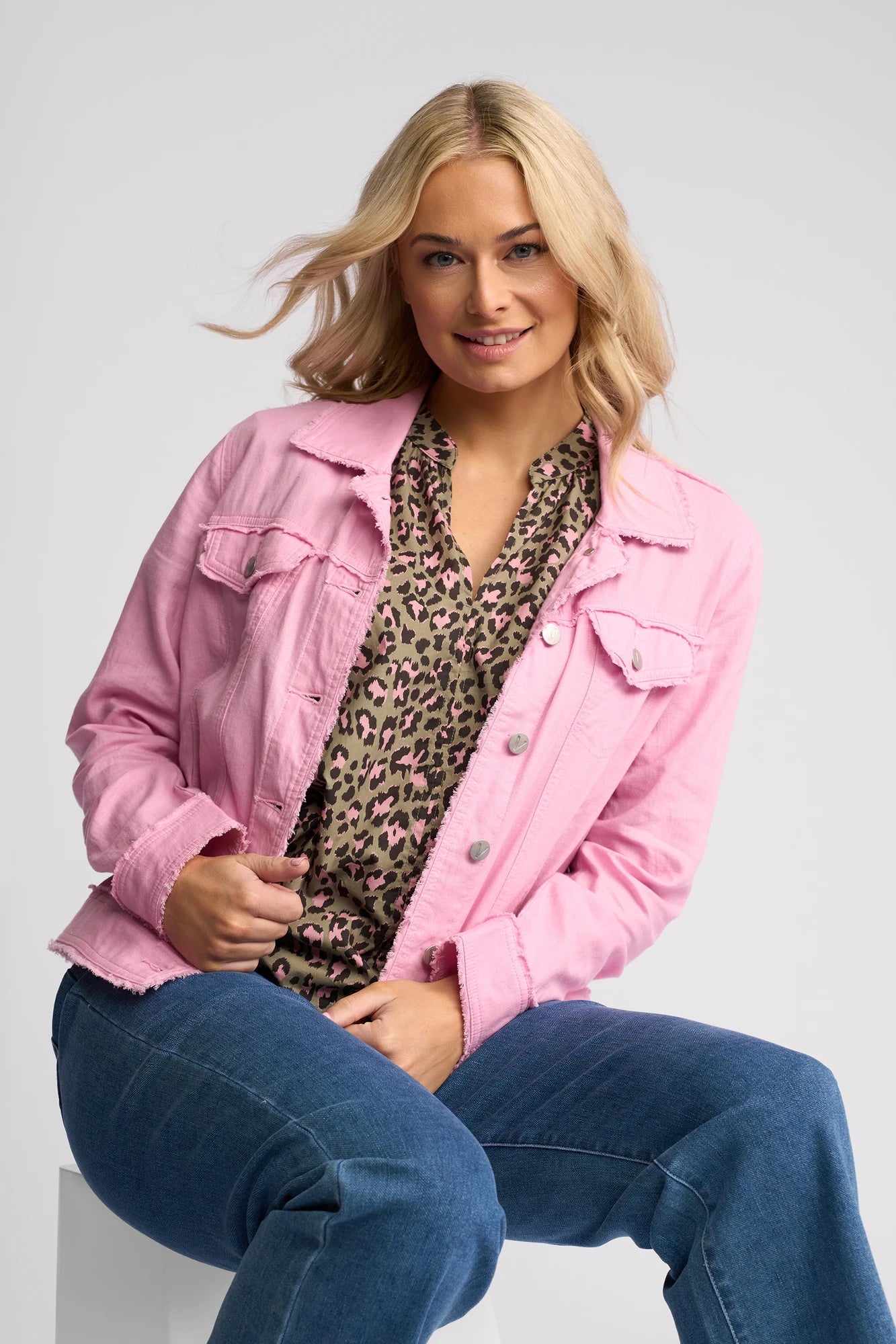 A woman with blonde hair sits on a stool, wearing the Vassalli Aura Jacket in Taffy—a light pink denim jacket—over a leopard print blouse and blue jeans, smiling at the camera against a light gray background.