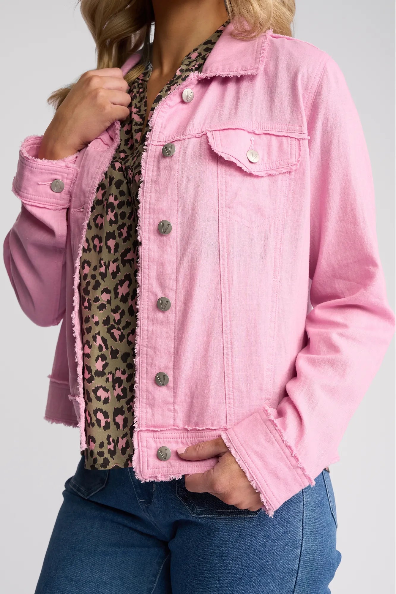 A person wearing a Vassalli Aura Jacket - Taffy layered over a leopard print blouse and styled with blue jeans. One hand is tucked into a pocket while the other lightly touches the collar, highlighting this versatile summer jacket.