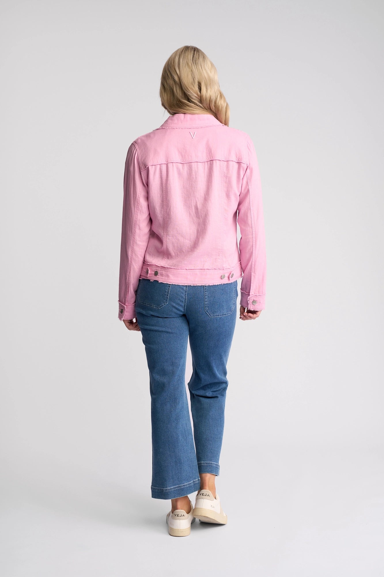 A person with blonde hair stands facing away, wearing the Vassalli Aura Jacket in Taffy, blue jeans, and white sneakers against a plain light background.