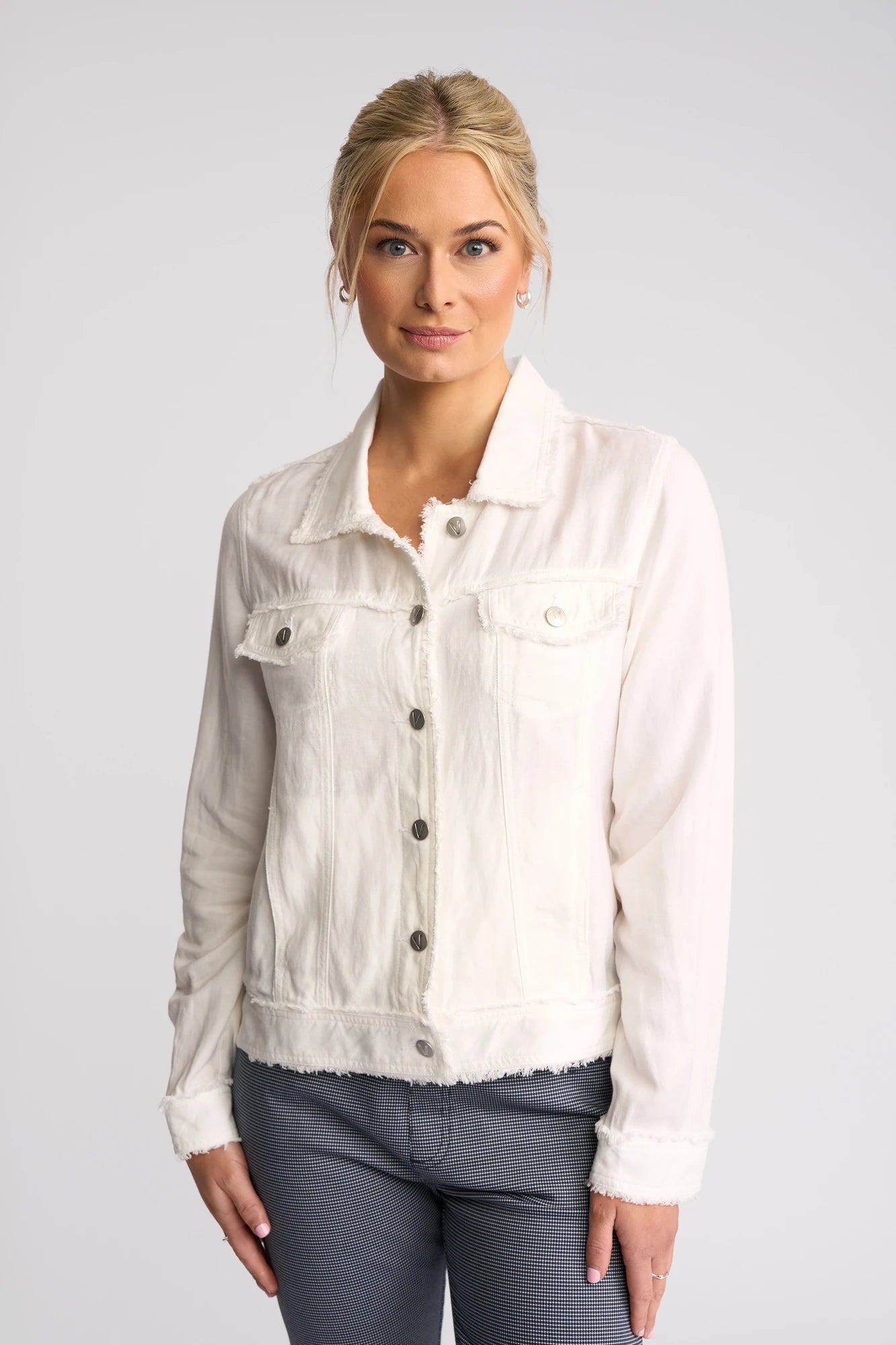 A woman with blonde hair in a low bun wears the Vassalli Aura Jacket - White, a button-up style crafted from a viscose linen blend, paired with gray pants. She stands against a plain light gray background, looking at the camera with a neutral expression.