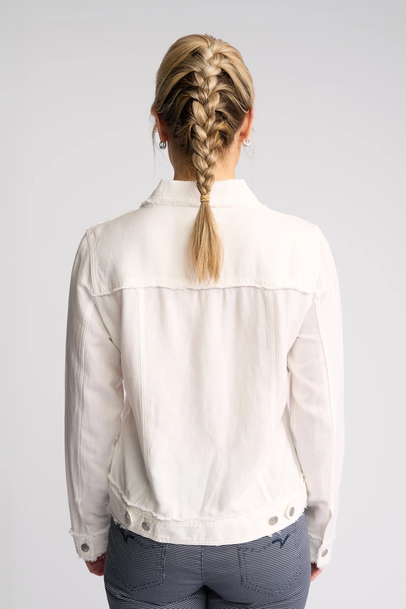 A person with blonde hair in a braid is shown from the back against a plain light background, wearing gray pants and the Vassalli Aura Jacket in white.