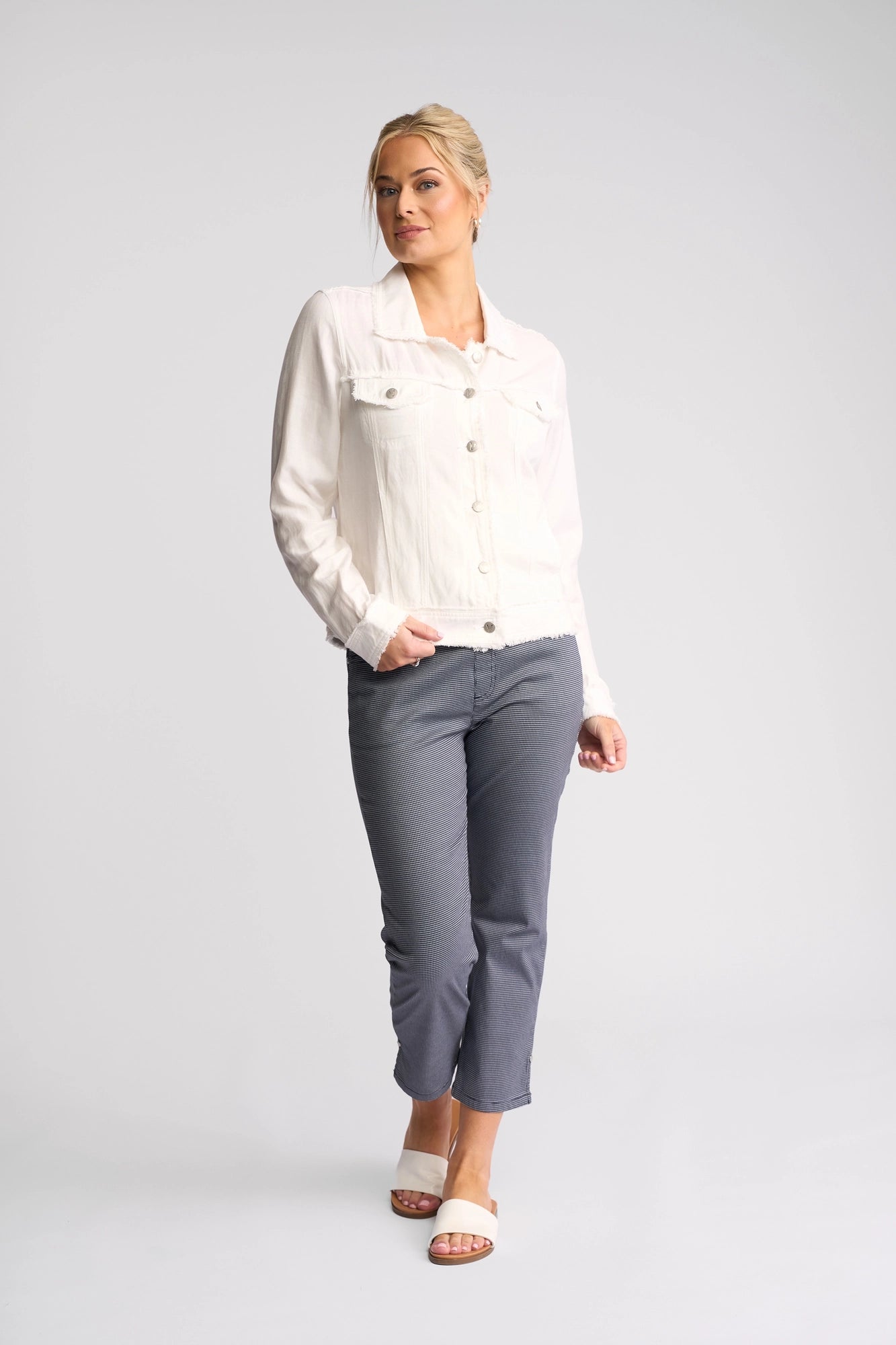 A woman with blonde hair pulled back stands against a plain background, wearing the Vassalli Aura Jacket in white. She pairs the crisp denim jacket with gray ankle-length pants and white slide sandals, looking slightly to the side with a relaxed expression.
