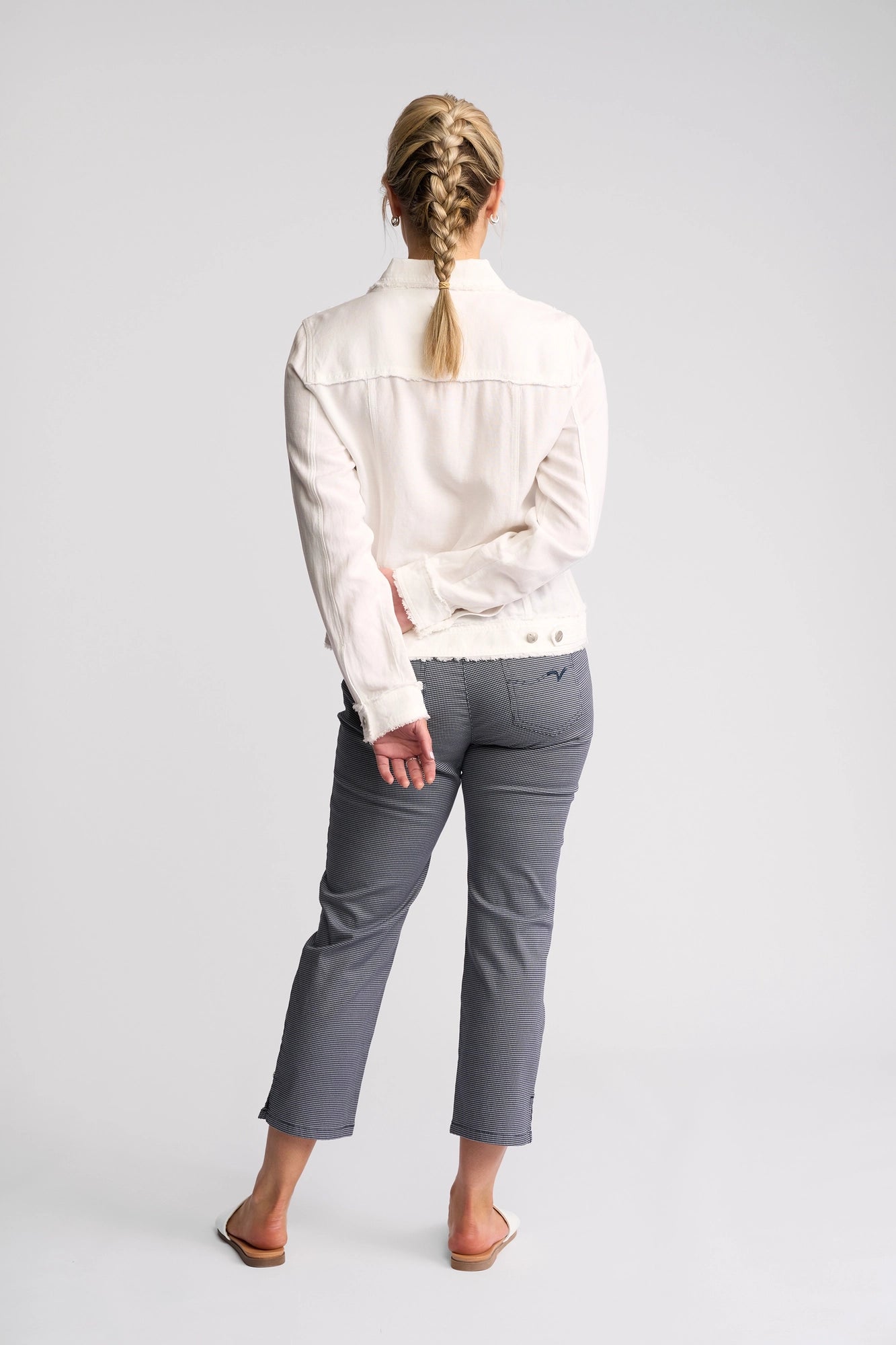 A person with blond hair in a braid stands facing away, wearing the Vassalli Aura Jacket in white, gray pants, and white flats against a plain white background.