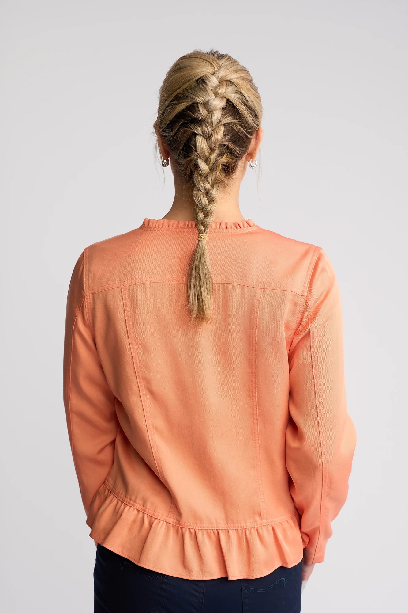 A person with blond hair styled in a French braid is seen from behind against a light gray background, wearing the Vassalli Sawyer Jacket in Apricot layered over a peach-colored long-sleeve blouse with ruffle details.