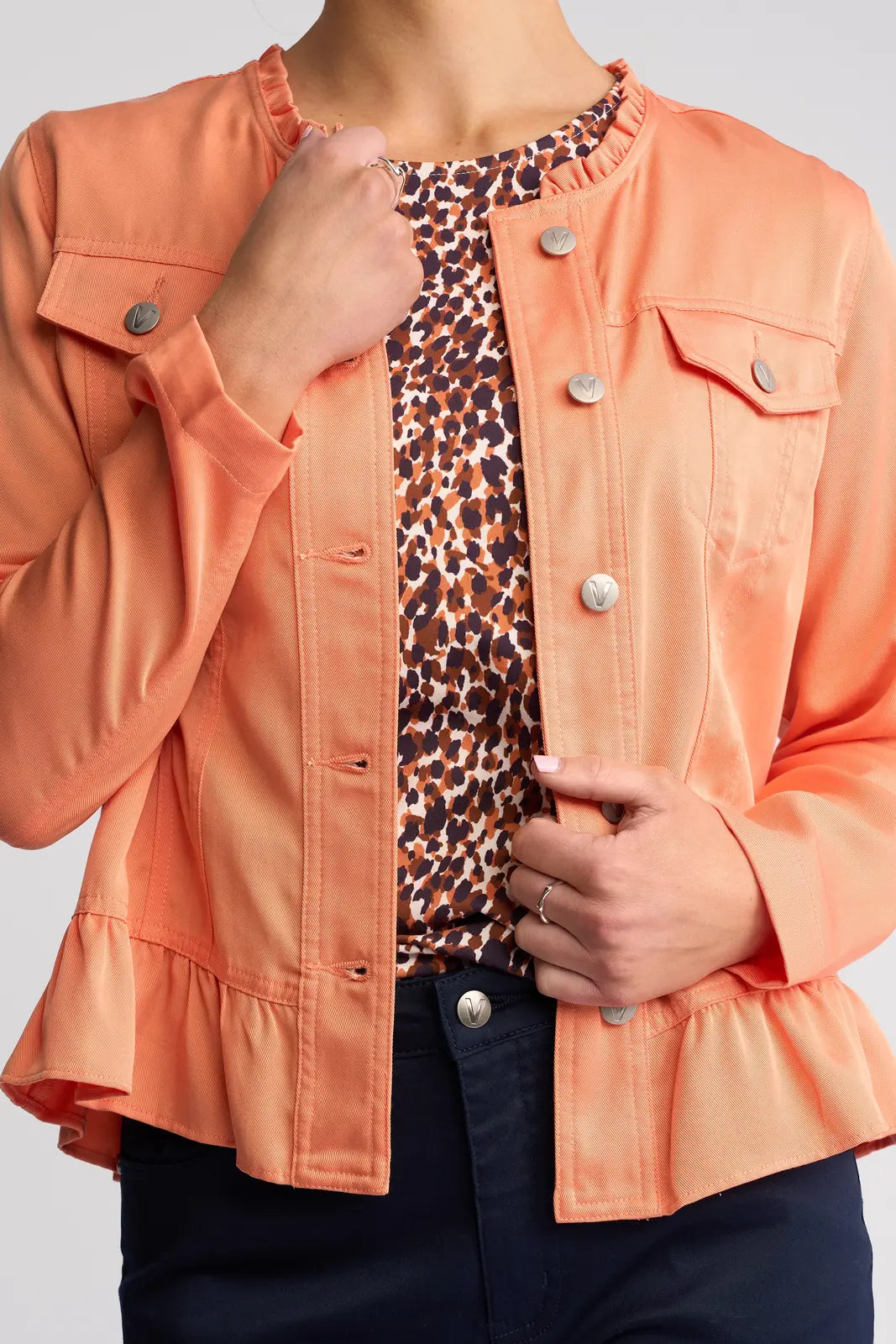 A woman is wearing the Vassalli Sawyer Jacket in Apricot—a lightweight Tencel piece featuring silver buttons—layered over a patterned shirt. She holds one lapel with one hand while her other hand rests near her waist, shown from shoulders to hips.