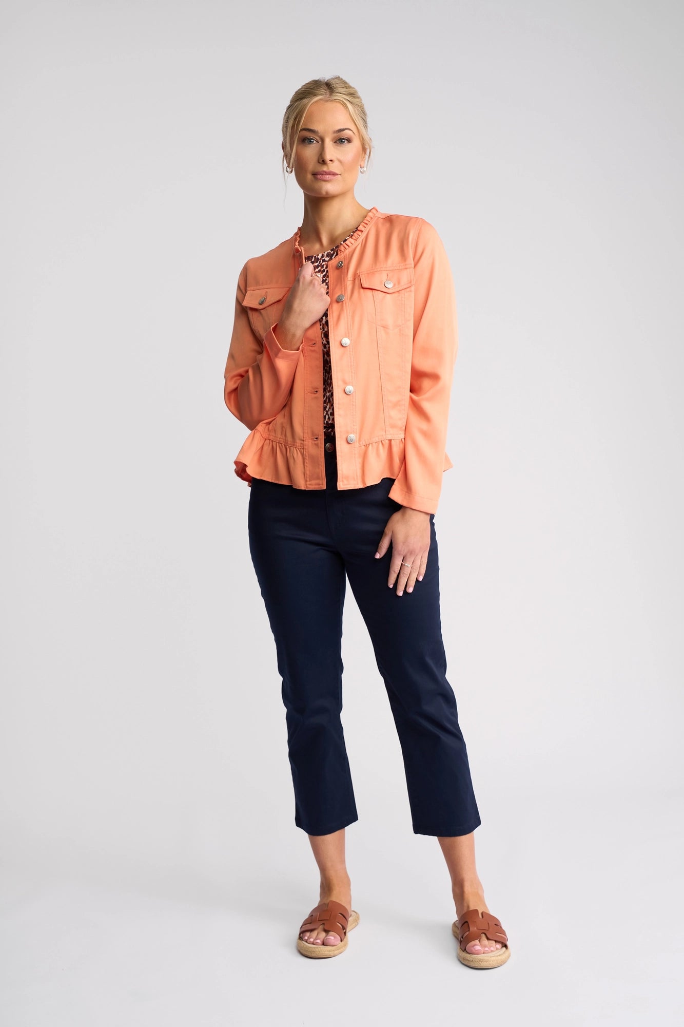 A woman stands against a plain background wearing the Vassalli Sawyer Jacket in Apricot, paired with a patterned top, navy pants, and sandals. She faces forward with one hand touching her chin and the other resting by her side.