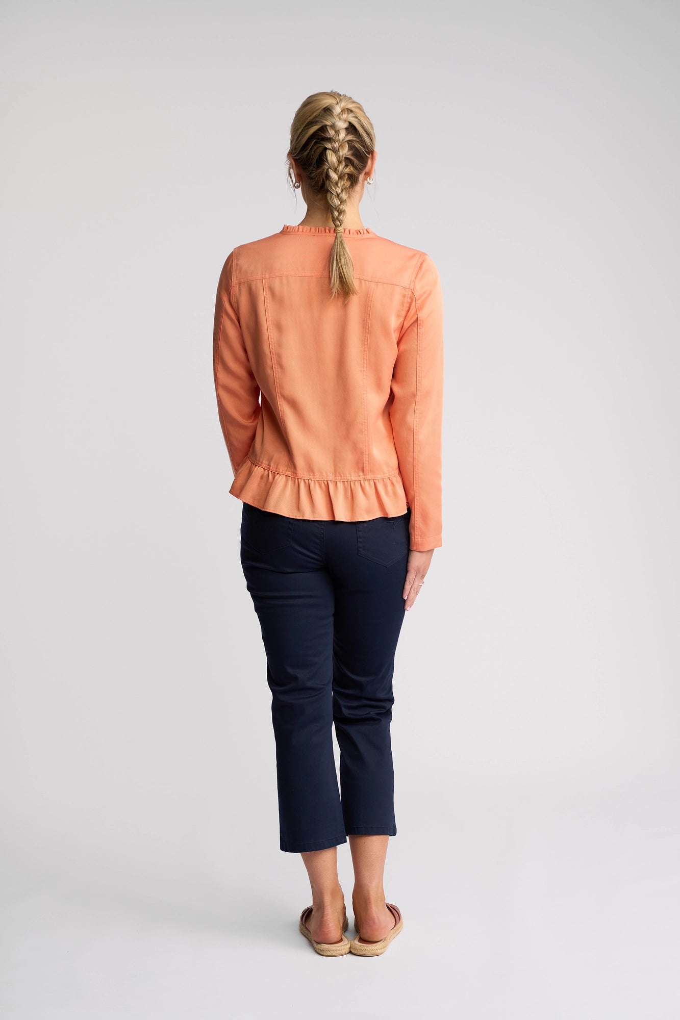 A woman with blond braided hair, dressed in the Vassalli Sawyer Jacket in Apricot, navy pants, and sandals, stands facing away from the camera against a plain light background.