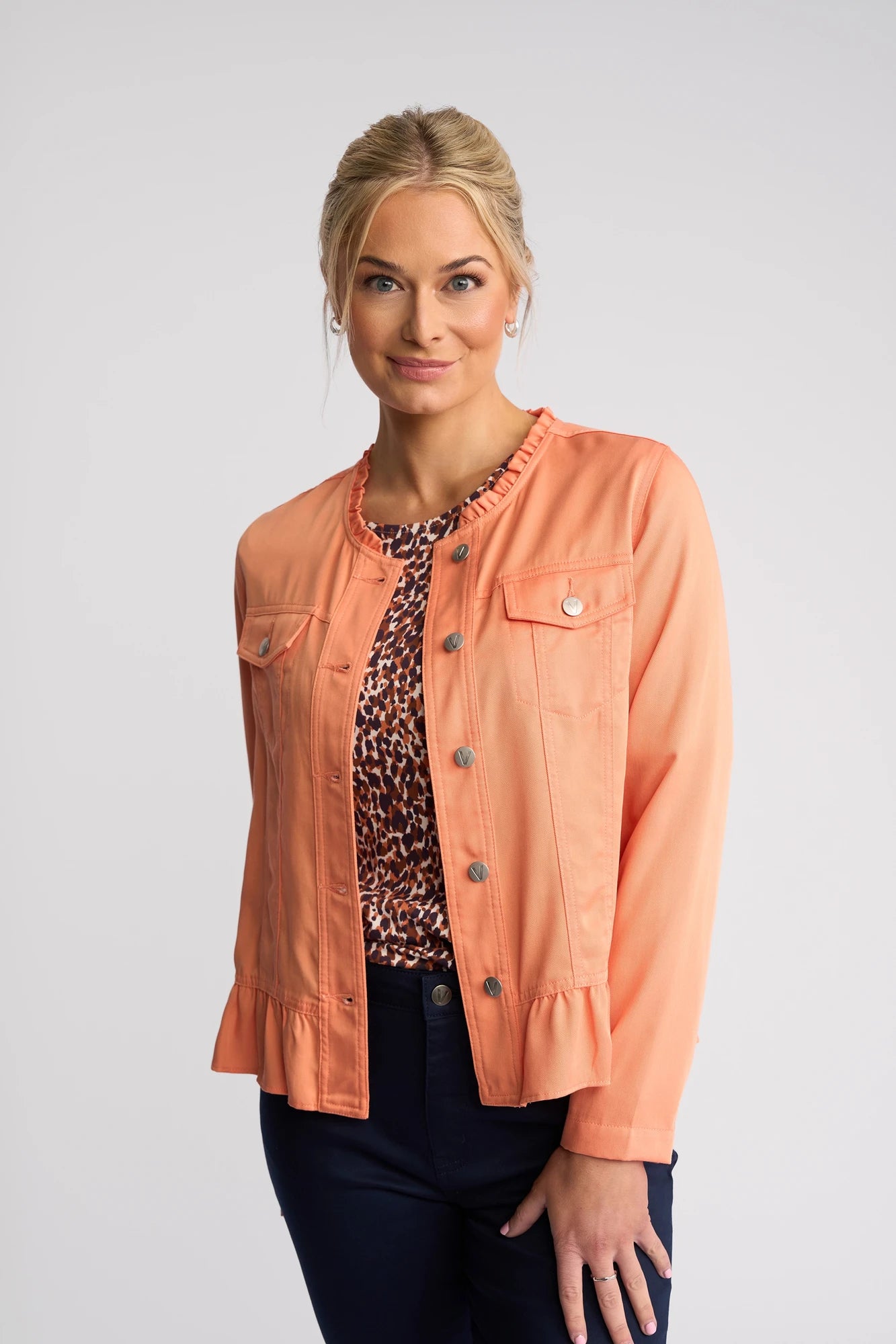 A woman with blonde hair in a low bun wears the Sawyer Jacket in Apricot by Vassalli—a stylish, collarless jacket crafted from lightweight tencel—over a leopard print top and dark pants. She stands against a plain light background, smiling softly at the camera.