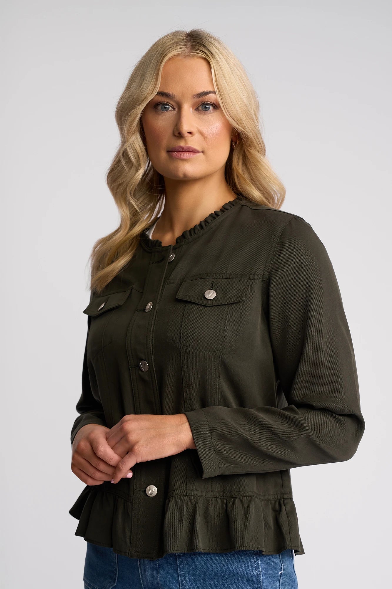 A woman with long blonde hair wearing a khaki Vassalli Sawyer Jacket, a stylish collarless jacket with ruffled trim, stands against a light gray background, looking slightly to the right.