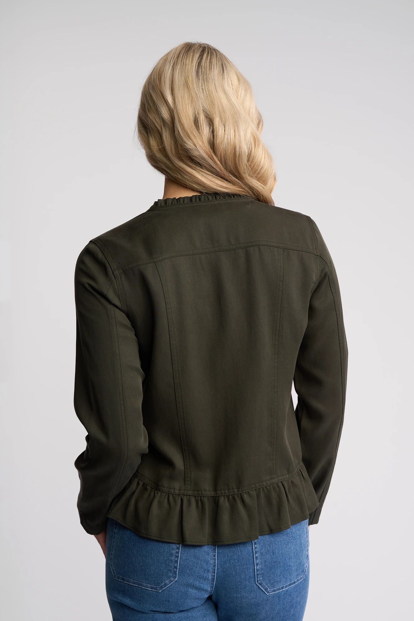 A person with long blonde hair is shown from the back, wearing a Vassalli Sawyer Jacket in Khaki with a ruffled hem and blue jeans against a plain light background.