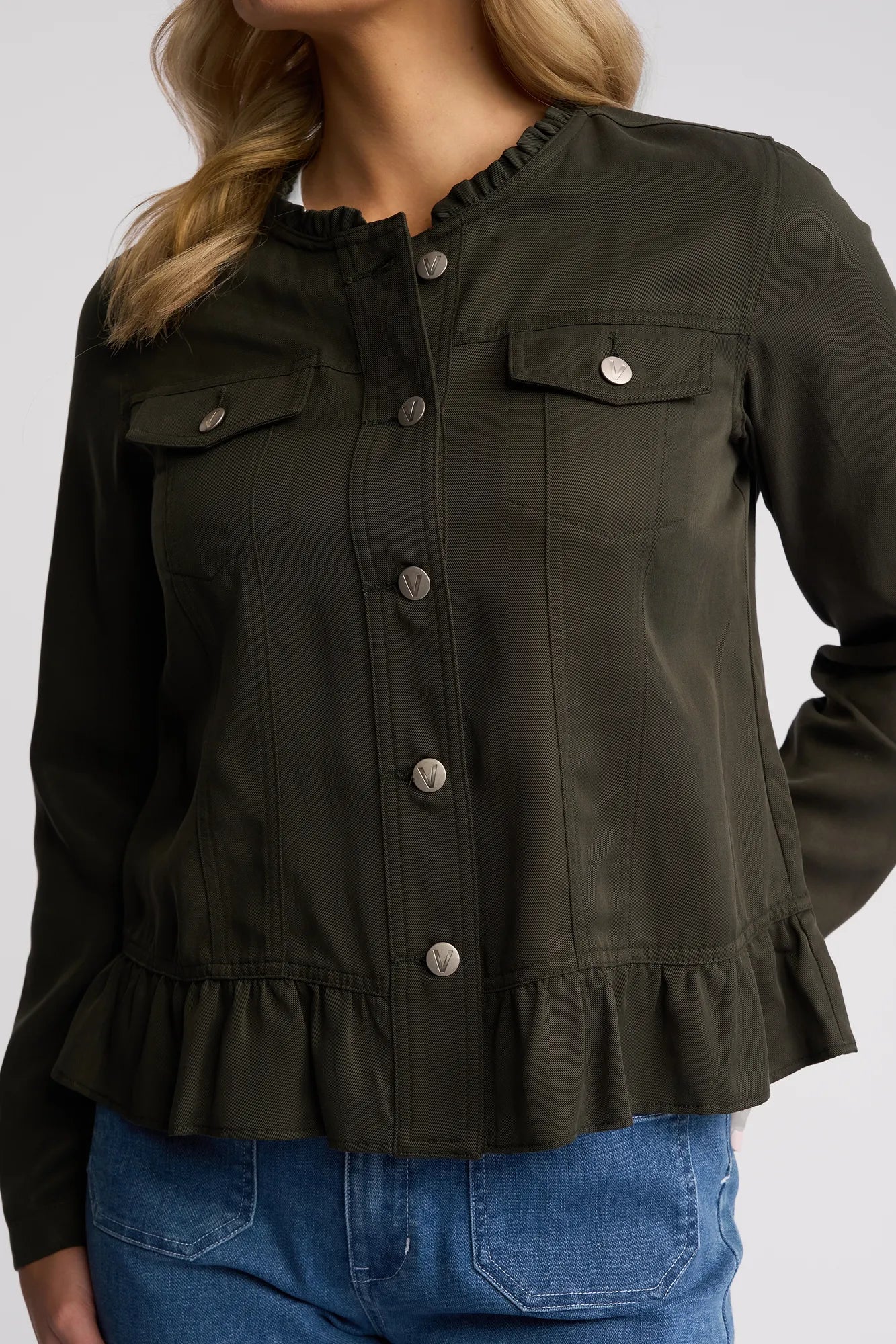 A woman wearing a khaki Vassalli Sawyer Jacket—a lightweight tencel, collarless jacket with ruffled trim at the waist and chest pockets—paired with blue jeans. Her head is partially out of frame.