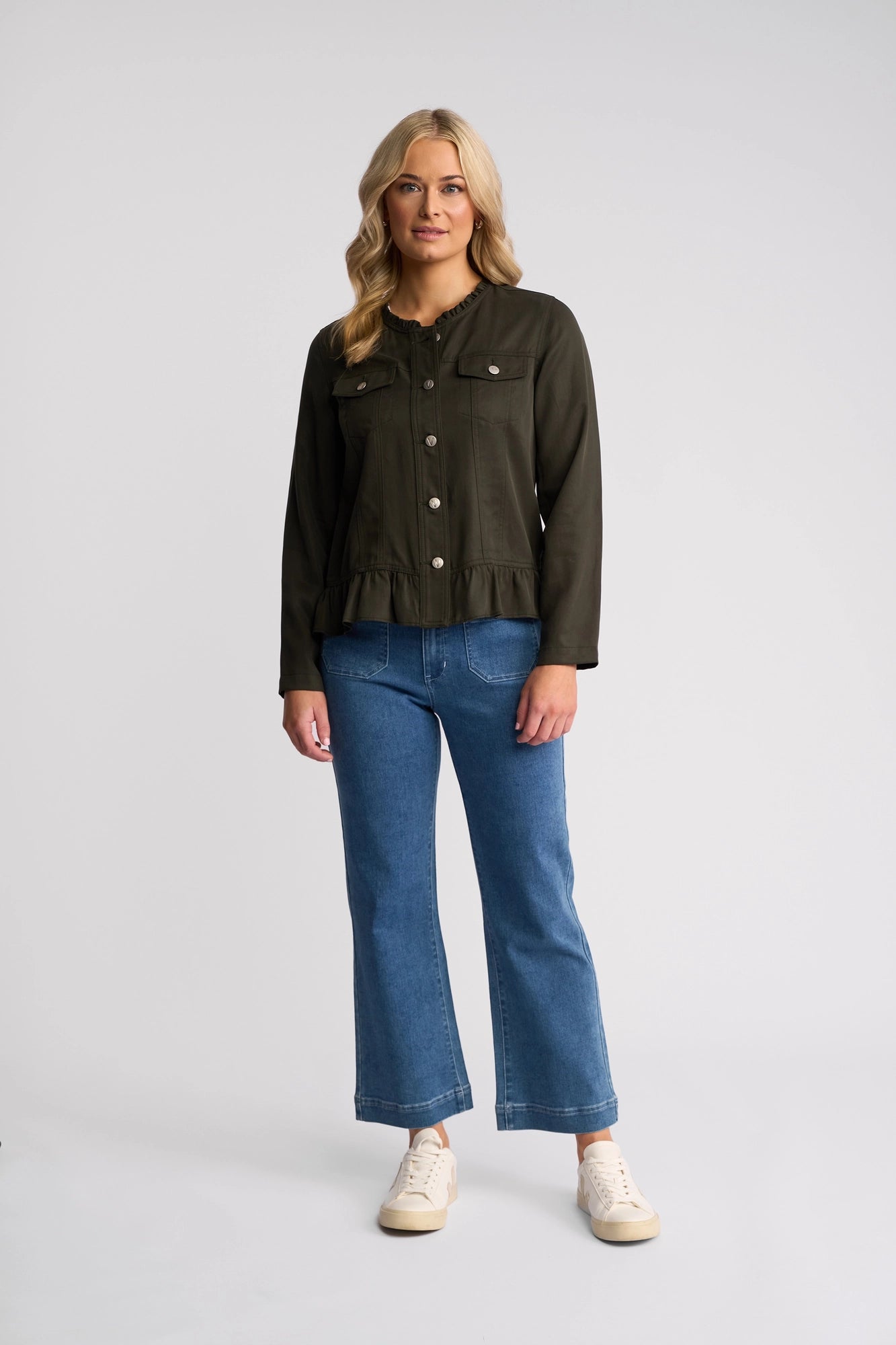 A woman stands against a plain background, wearing the Vassalli Sawyer Jacket in Khaki—a lightweight Tencel jacket with a modern, collarless design—paired with blue jeans and white sneakers. She has long blonde hair and a neutral expression.