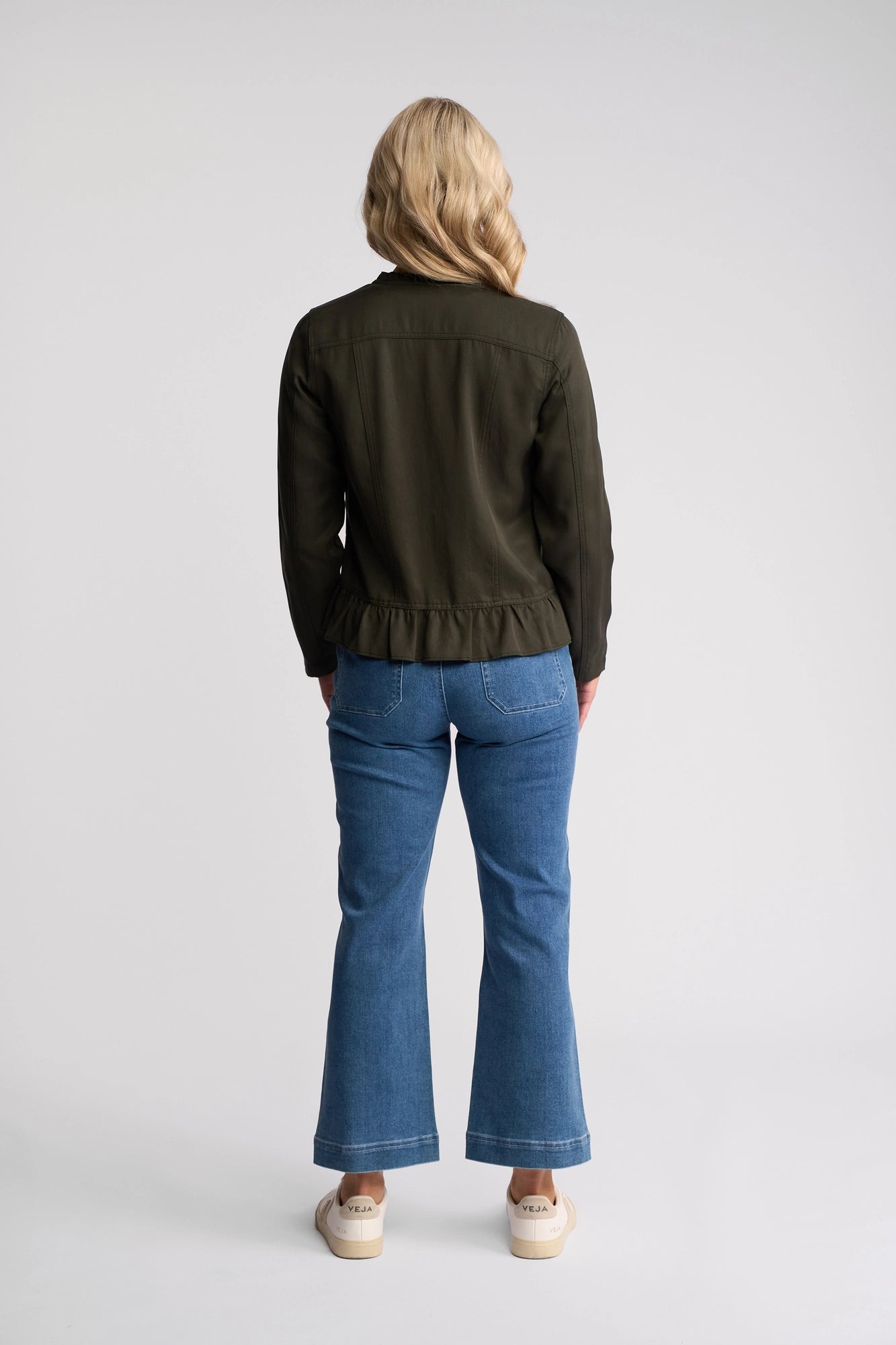 A person with blonde hair is seen from behind, wearing a khaki Sawyer Jacket by Vassalli, blue jeans, and light-colored shoes against a plain, light background.