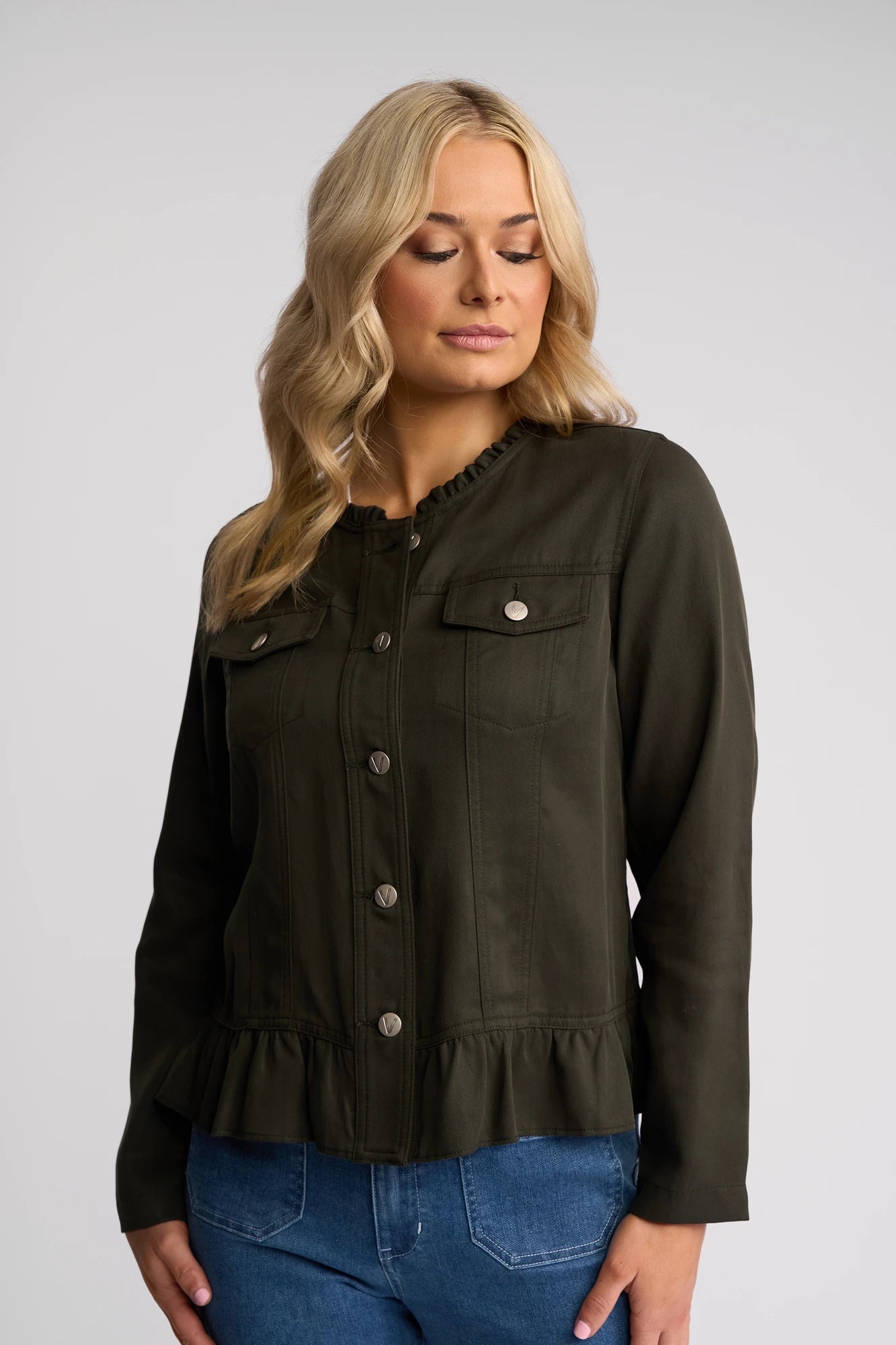 A woman with long blonde hair wears the Vassalli Sawyer Jacket in Khaki—a stylish, collarless jacket with ruffle detail at the hem—paired with blue jeans, standing against a plain light gray background.