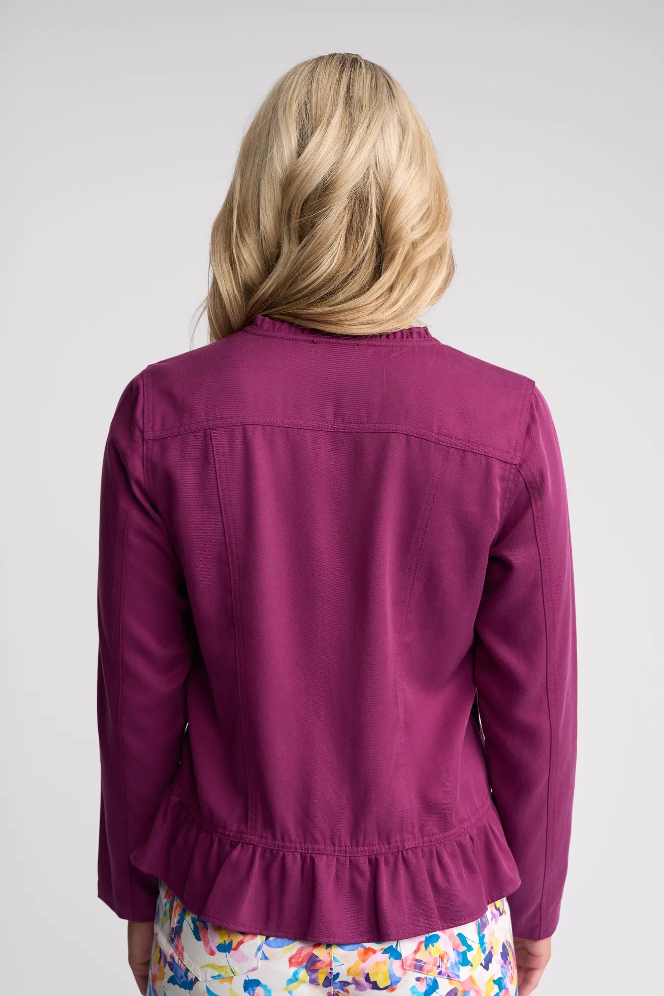 A person with blonde hair, seen from behind, is wearing a vibrant plum Vassalli Sawyer Jacket with a ruffled hem and a colorful, patterned skirt against a plain light background.