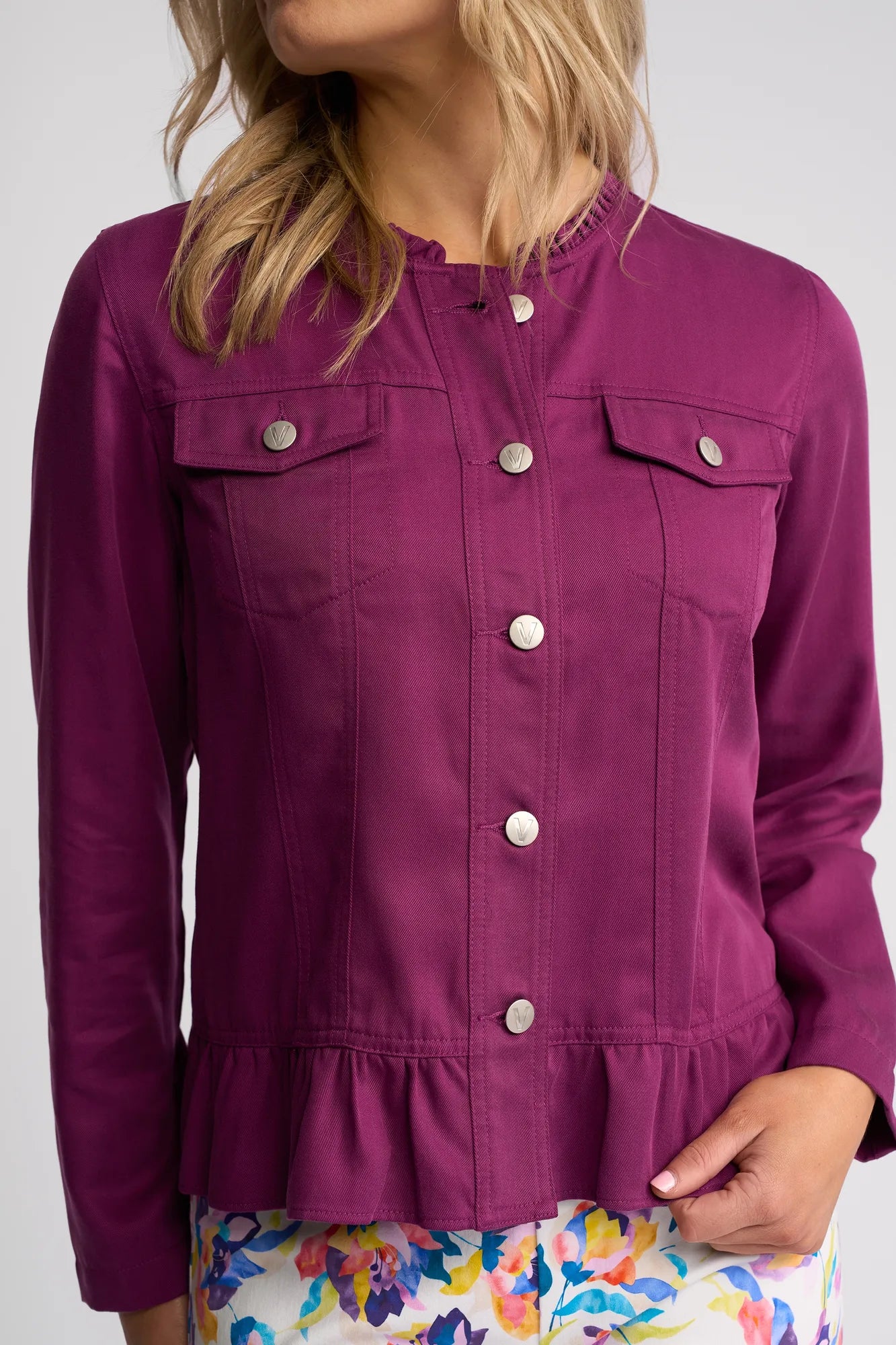 A woman wears a Vassalli Sawyer Jacket in Plum, a collarless, lightweight tencel jacket with front pockets and a ruffled hem, paired with a colorful floral skirt against a plain background.