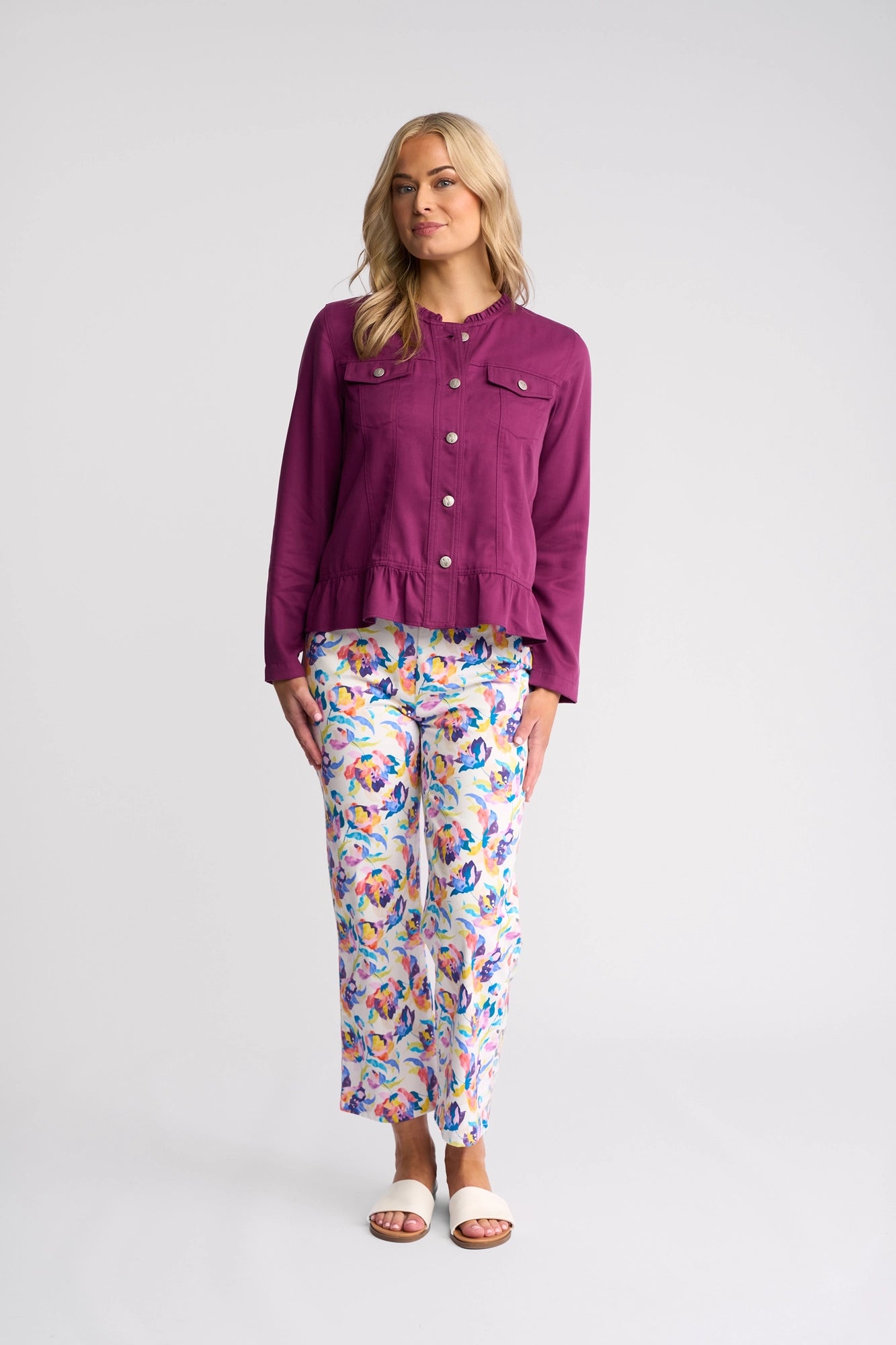 A woman with blonde hair stands against a light background, wearing the Vassalli Sawyer Jacket in Plum—a lightweight, collarless tencel jacket—styled with floral print pants in vibrant colors and white sandals. She has a relaxed expression and her hands rest at her sides.
