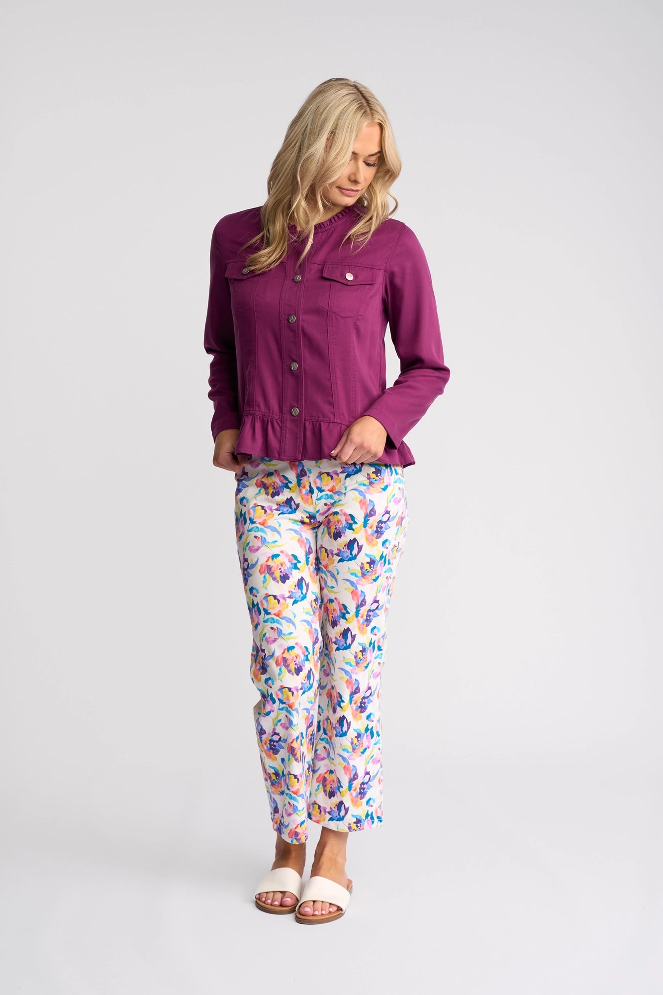 A woman stands looking down, wearing the Vassalli Sawyer Jacket in Plum, paired with colorful floral print pants and white sandals, against a plain white background.