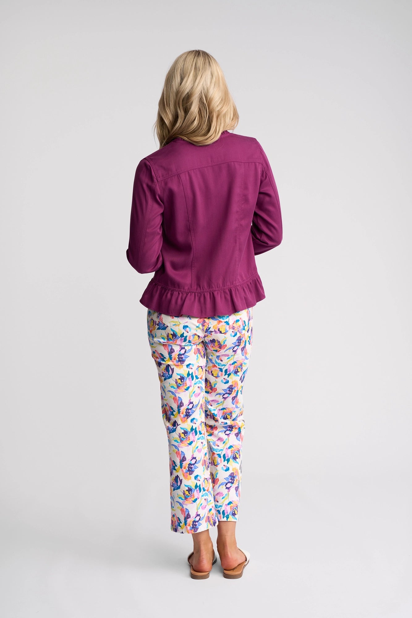 A woman with blonde hair stands facing away, wearing the Vassalli Sawyer Jacket in Plum and white pants with a colorful floral pattern. The lightweight, collarless jacket offers effortless style. She is barefoot on a plain white background.