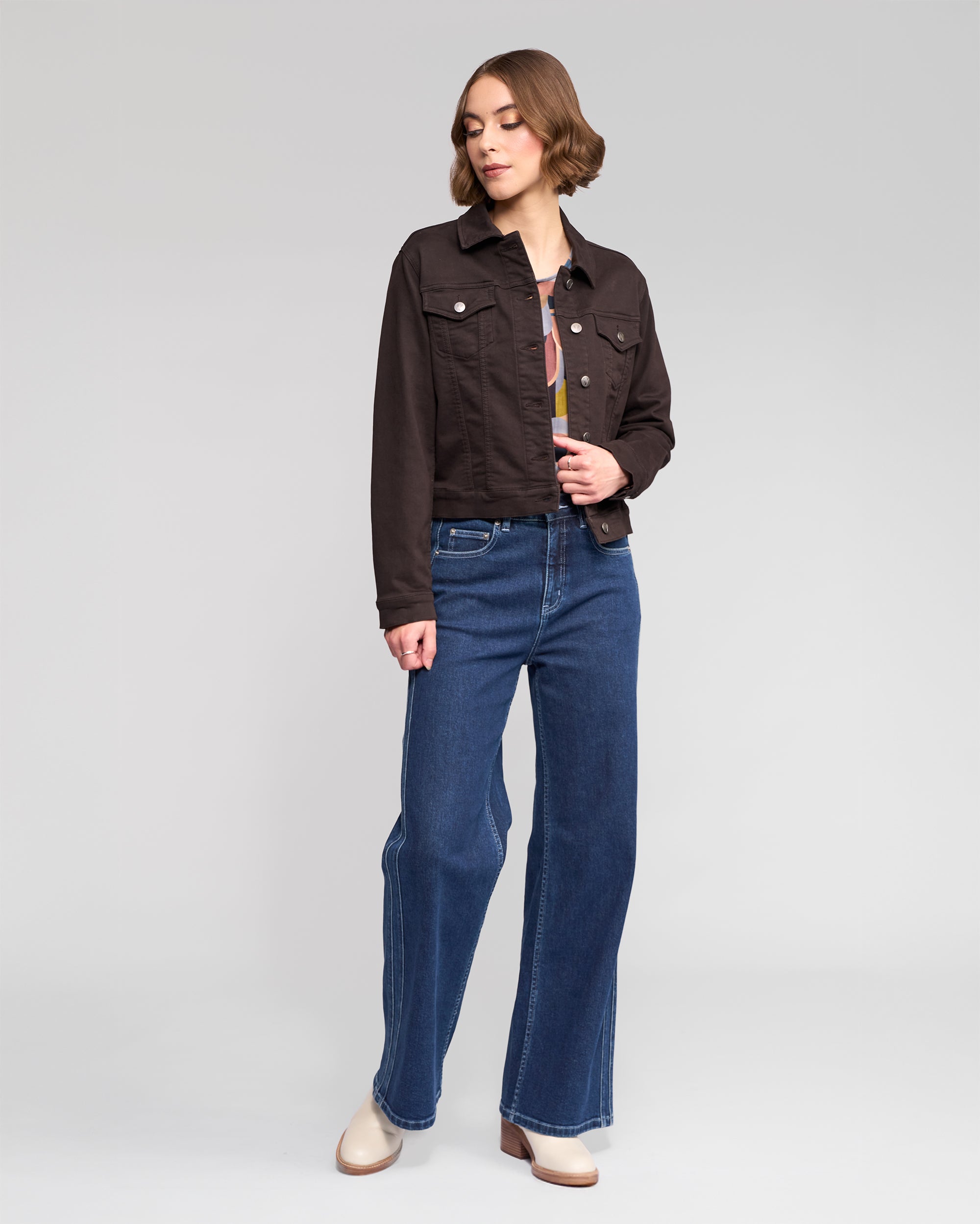 A woman stands against a plain background wearing the Vassalli Rider Knit Jacket in chocolate over a top, paired with blue high-waisted wide-leg jeans and beige shoes. She looks slightly to the side with one hand in her pocket.
