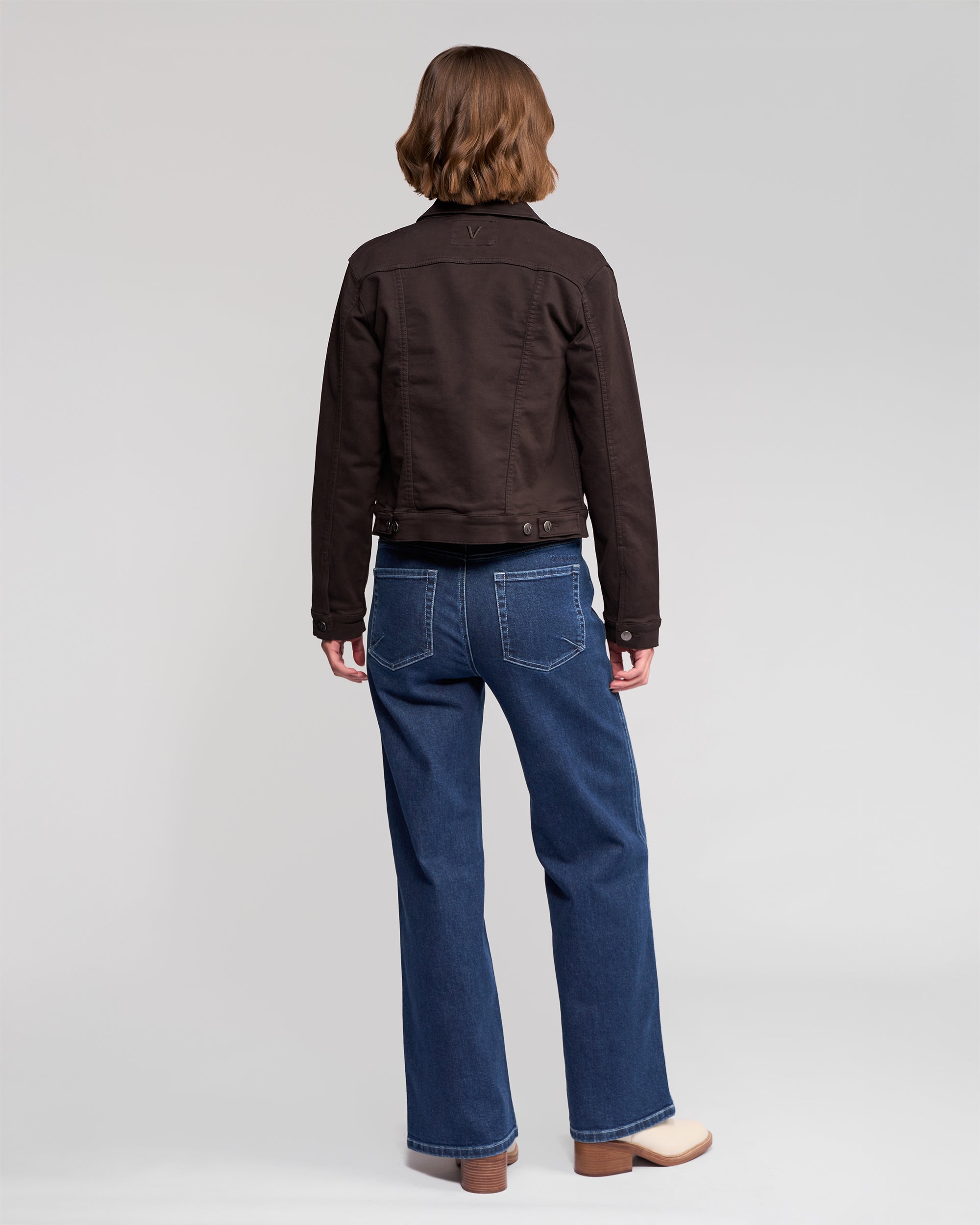 A person with short, wavy brown hair stands with their back to the camera, wearing the Vassalli Rider Knit Jacket in Chocolate, blue jeans, and beige shoes against a plain light grey background.