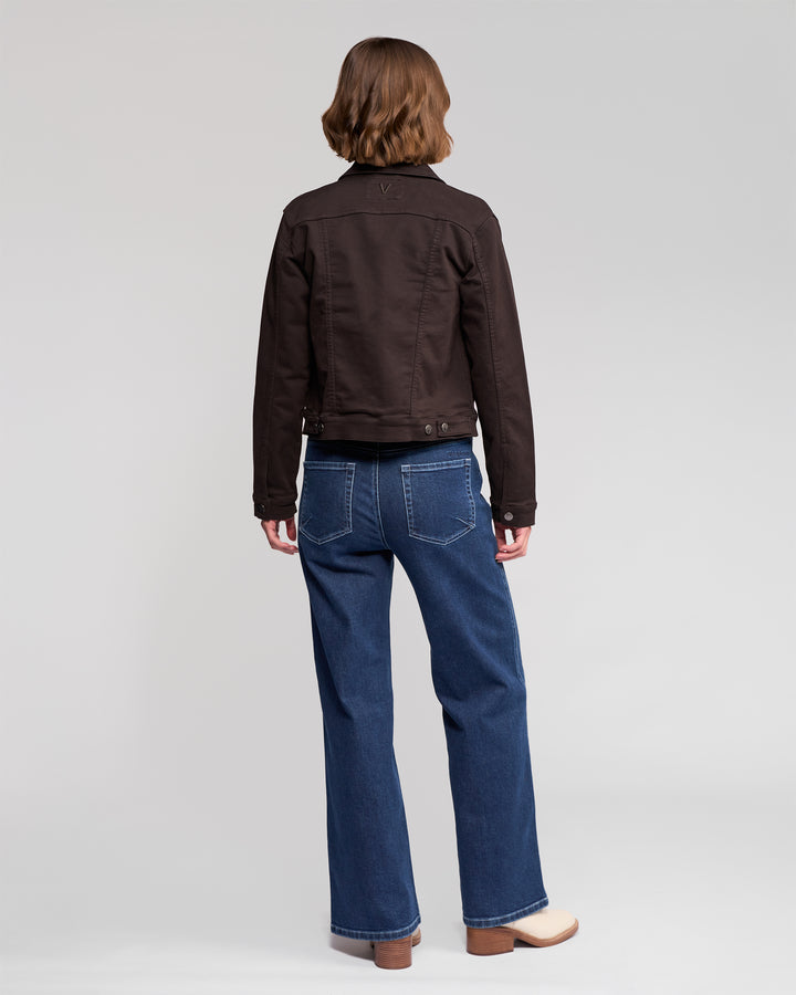 A person with short, wavy brown hair stands with their back to the camera, wearing the Vassalli Rider Knit Jacket in Chocolate, blue jeans, and beige shoes against a plain light grey background.