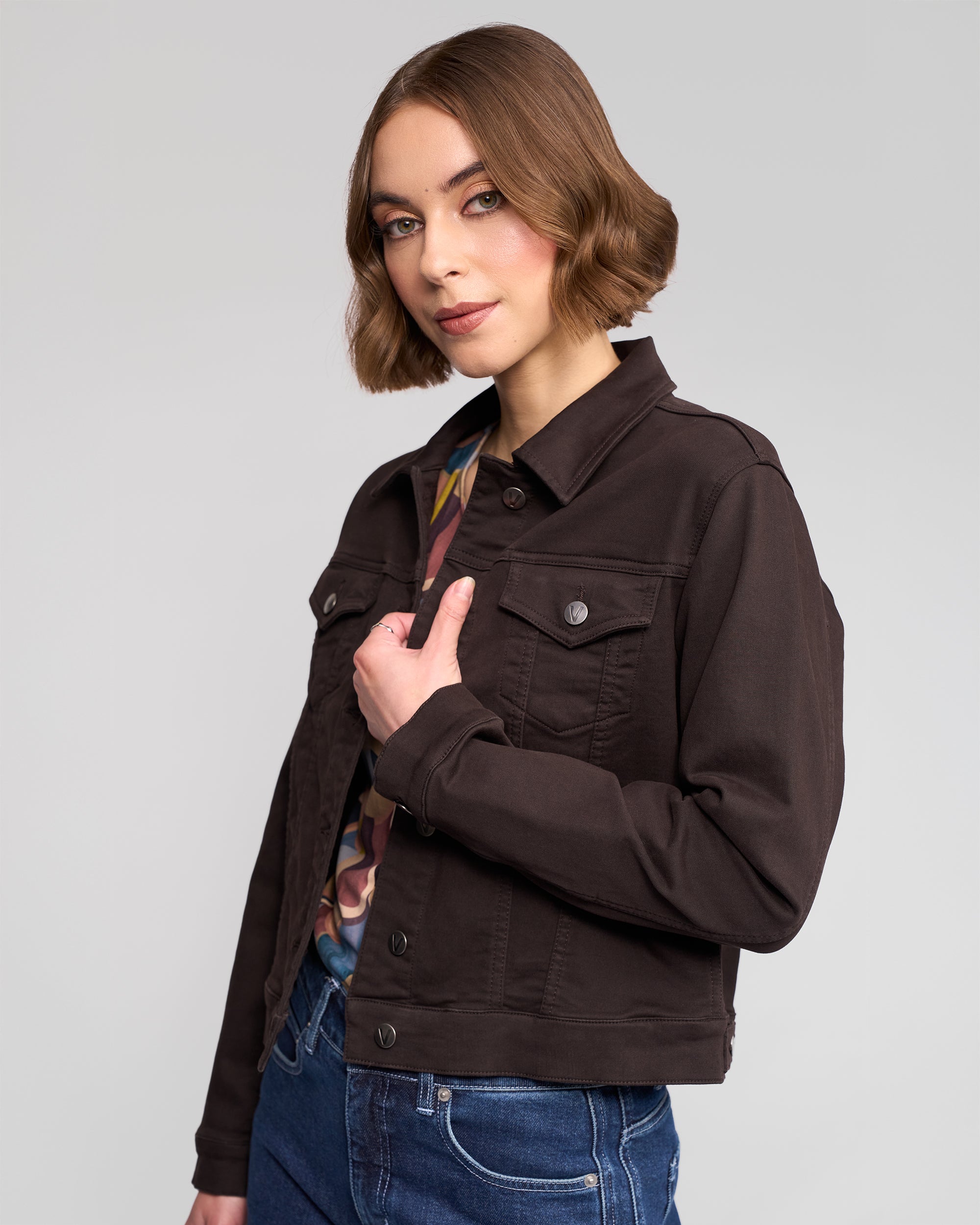 A woman with short brown hair models the Vassalli Rider Knit Jacket in Chocolate over a patterned top and blue jeans, posing confidently against a plain light gray background.