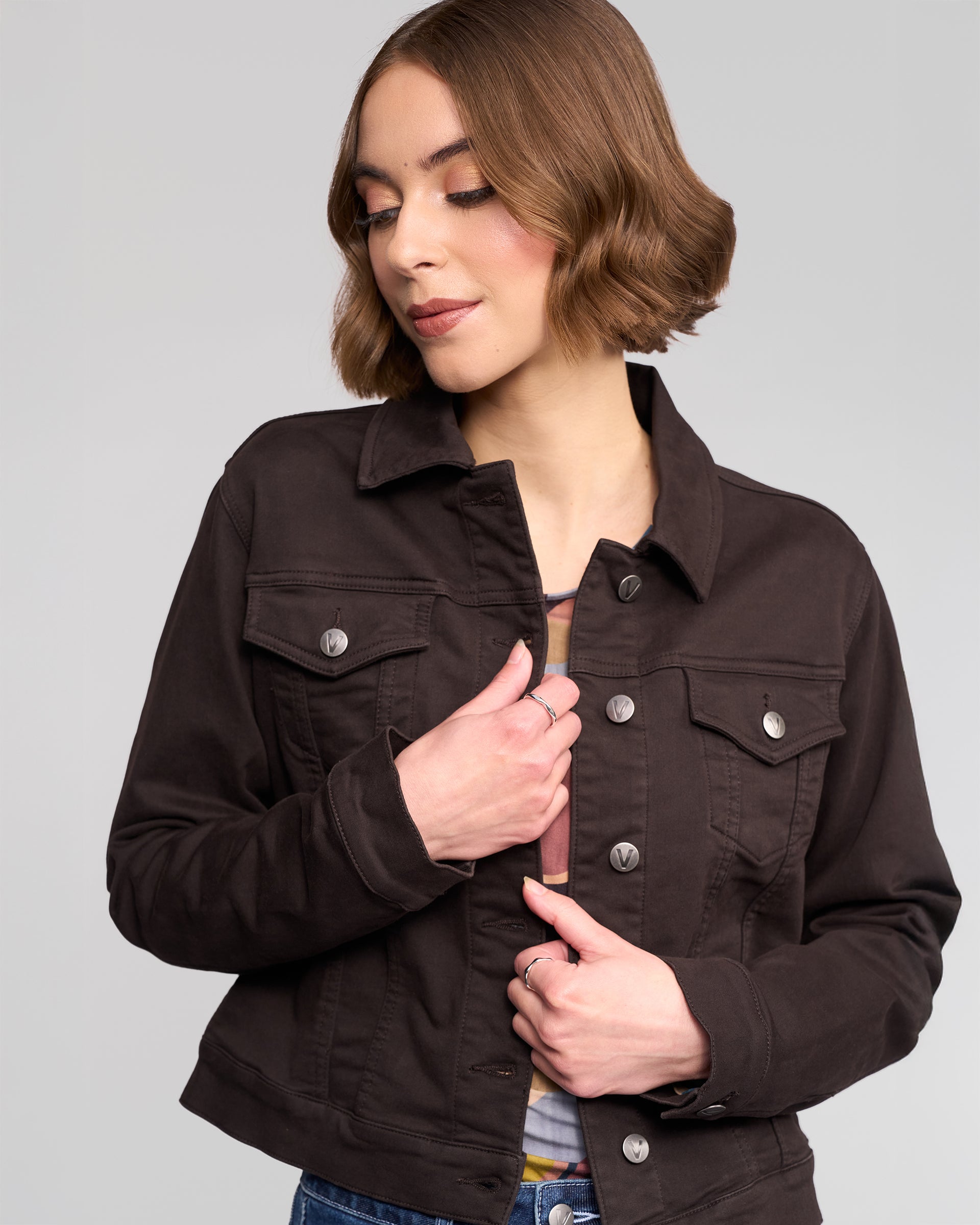 A woman with short brown hair wears the Vassalli Rider Knit Jacket in chocolate over a patterned shirt, looking down with a slight smile against a plain light gray background.