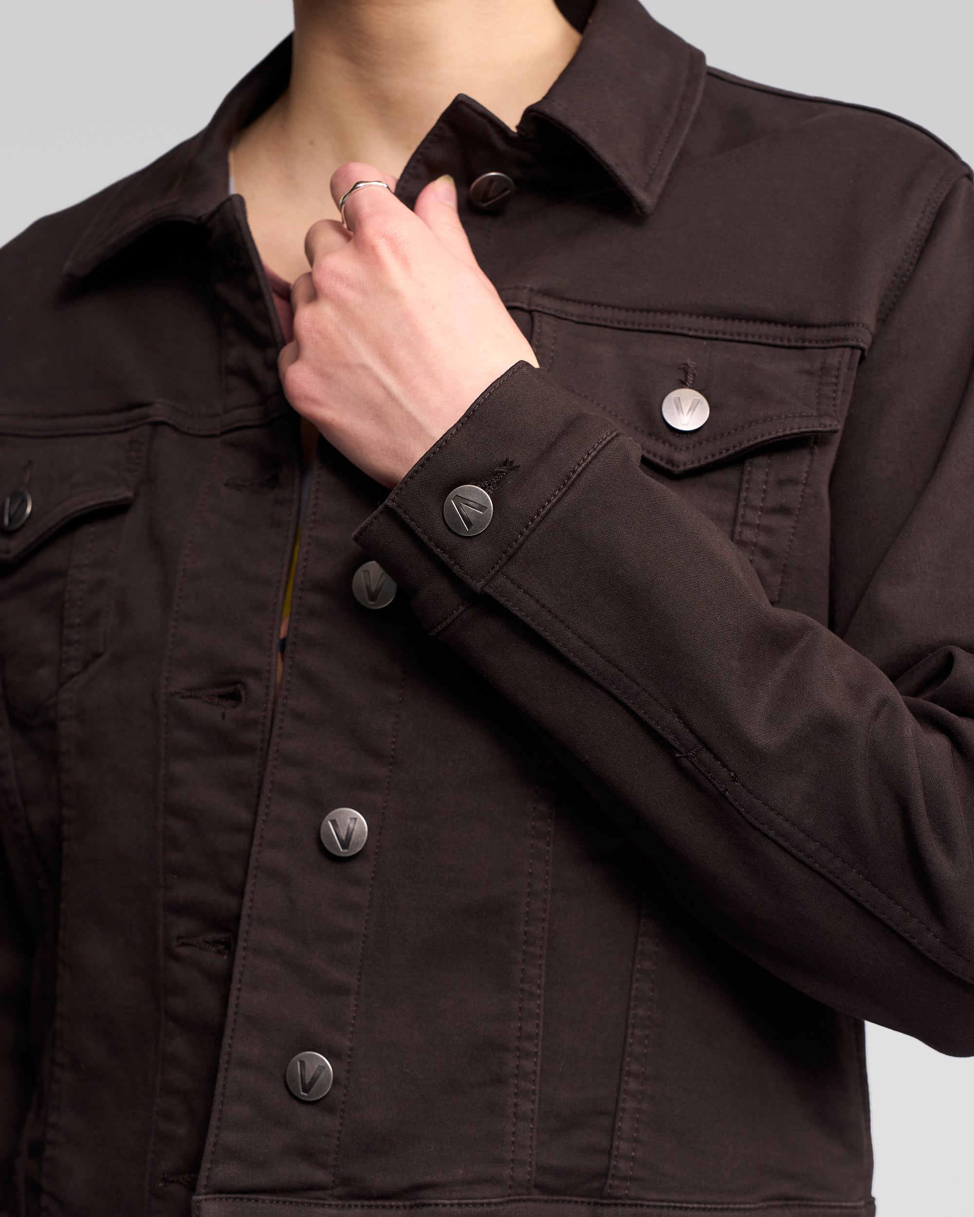 A person is holding the collar of the Vassalli Rider Knit Jacket - Chocolate, a relaxed fit style with metal buttons. The image is cropped to show only the torso and hand.