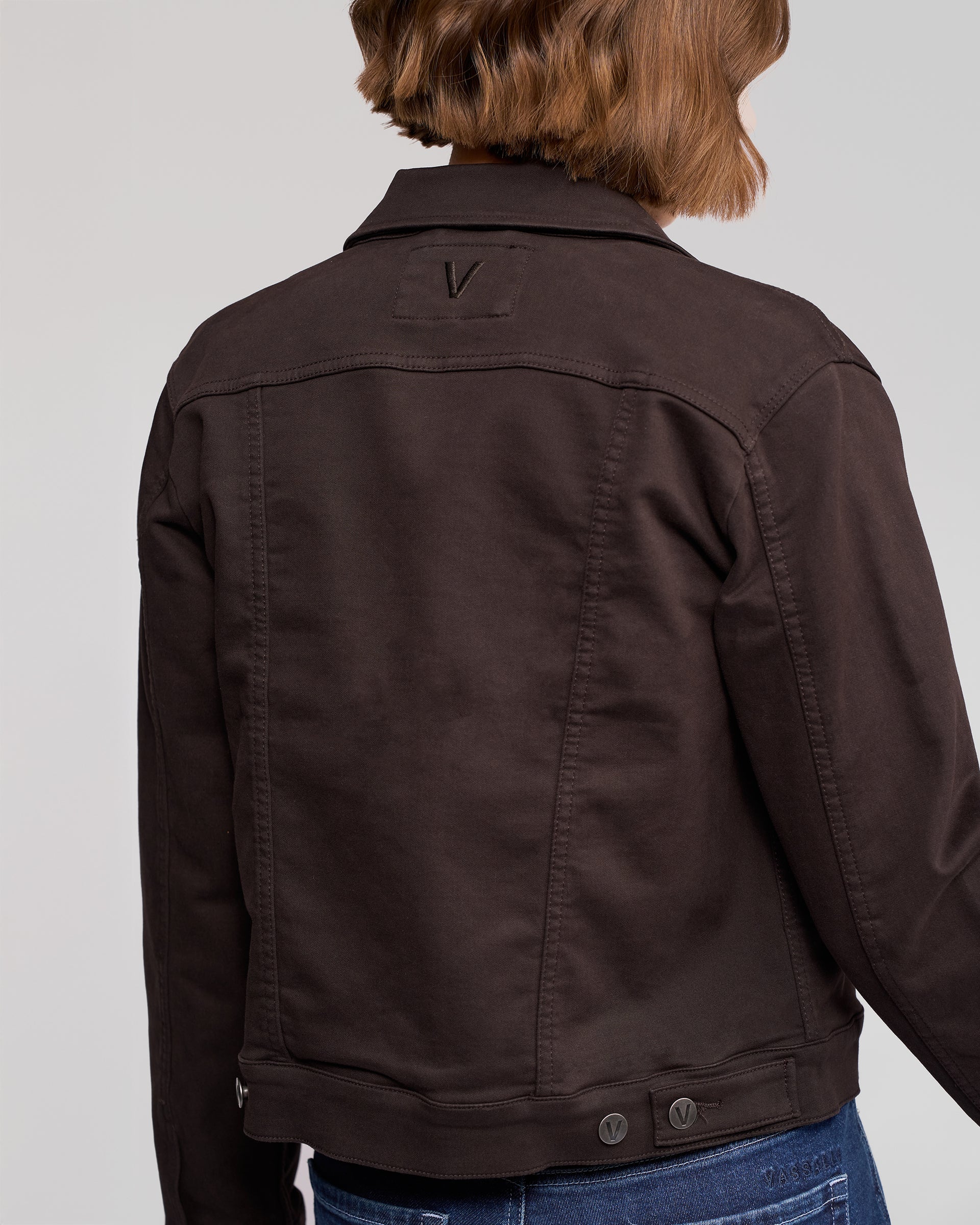 A person with wavy brown hair is shown from the back, wearing the Vassalli Rider Knit Jacket in chocolate brown with visible seams, buttons, a V detail on the upper back, and partially visible blue jeans.
